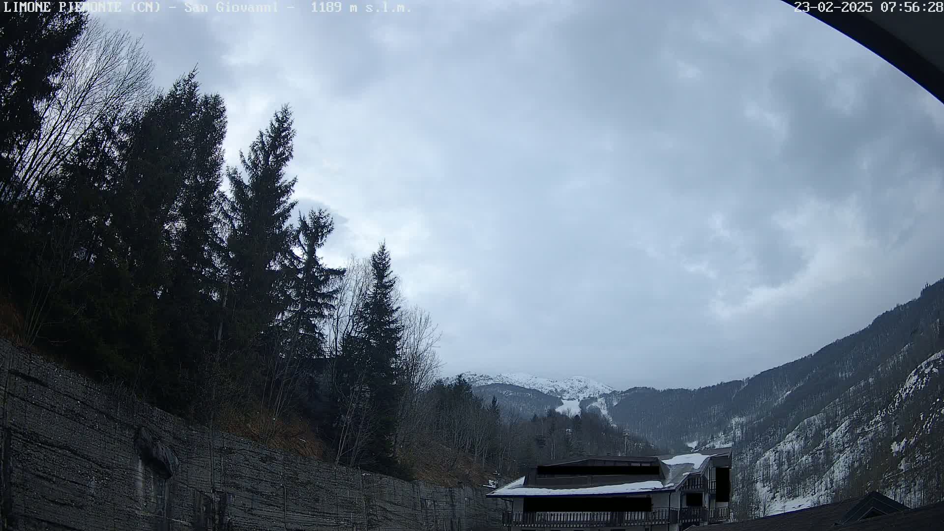 A cloudy day reveals a snow-dusted mountain range behind a forest of dark evergreens and a stone retaining wall, with a building partially visible in the foreground.