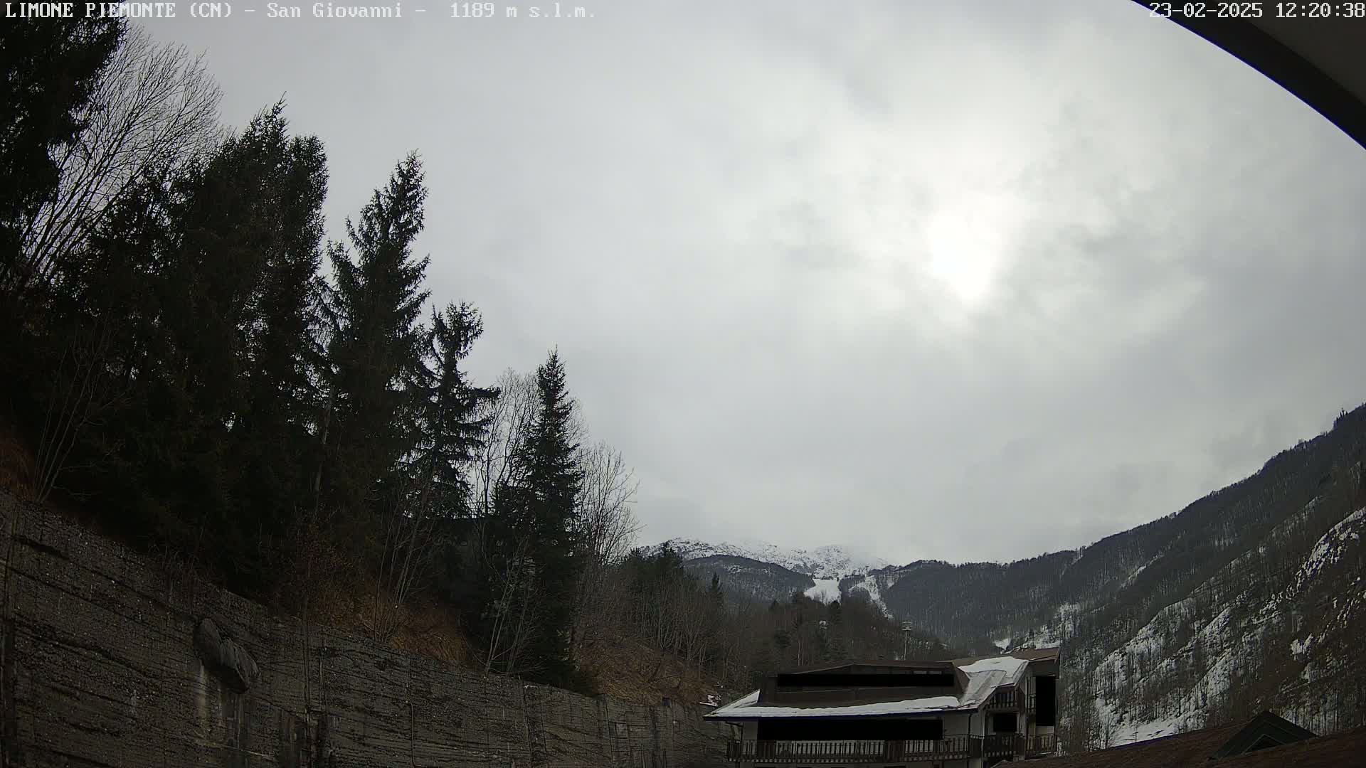A cloudy day reveals a mountainous landscape with snow-capped peaks in the distance, partially obscured by evergreens and a building nestled below.