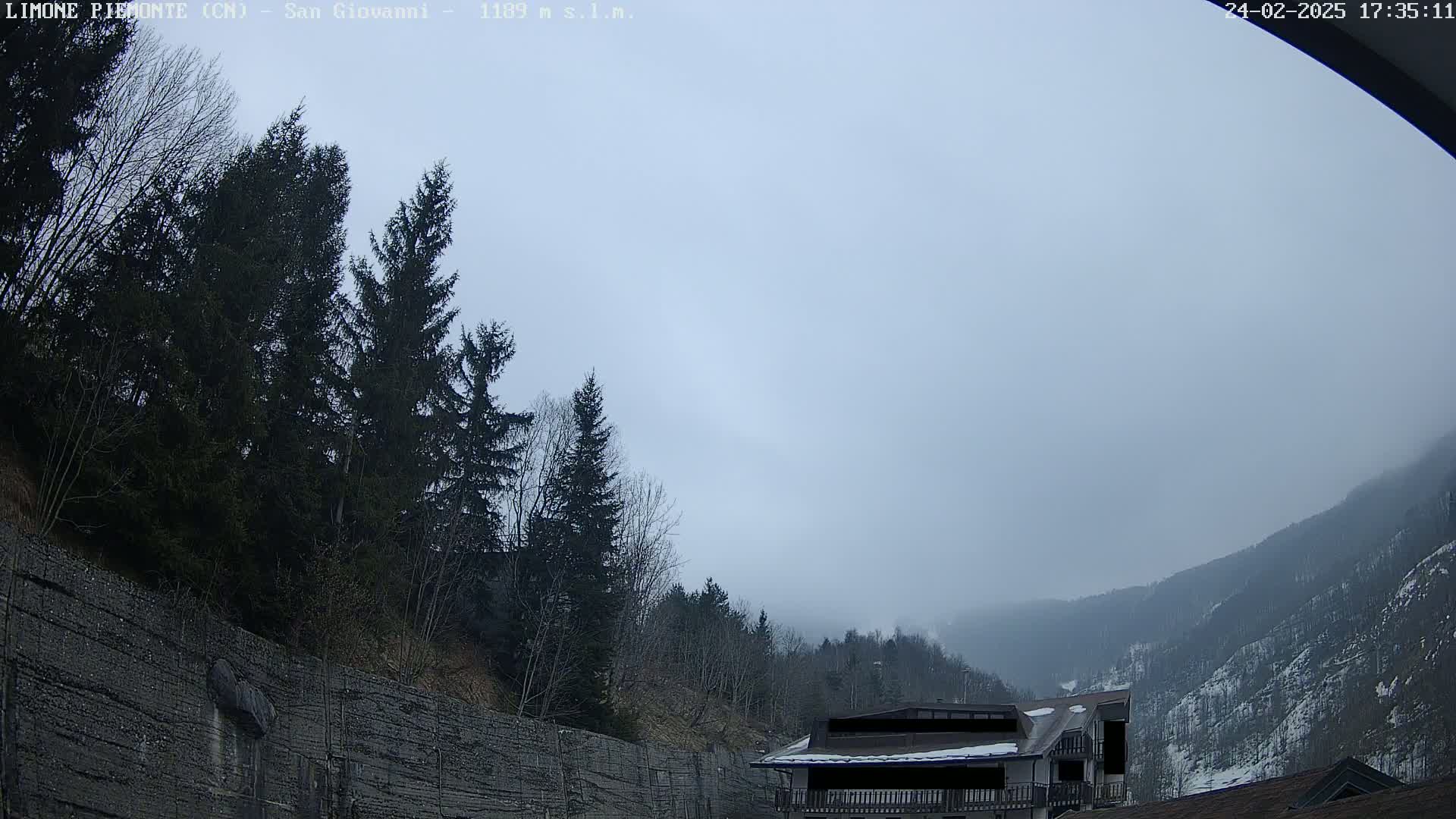 A hazy, overcast day reveals a mountain valley scene with evergreen trees, a stone retaining wall, and a partially visible building.