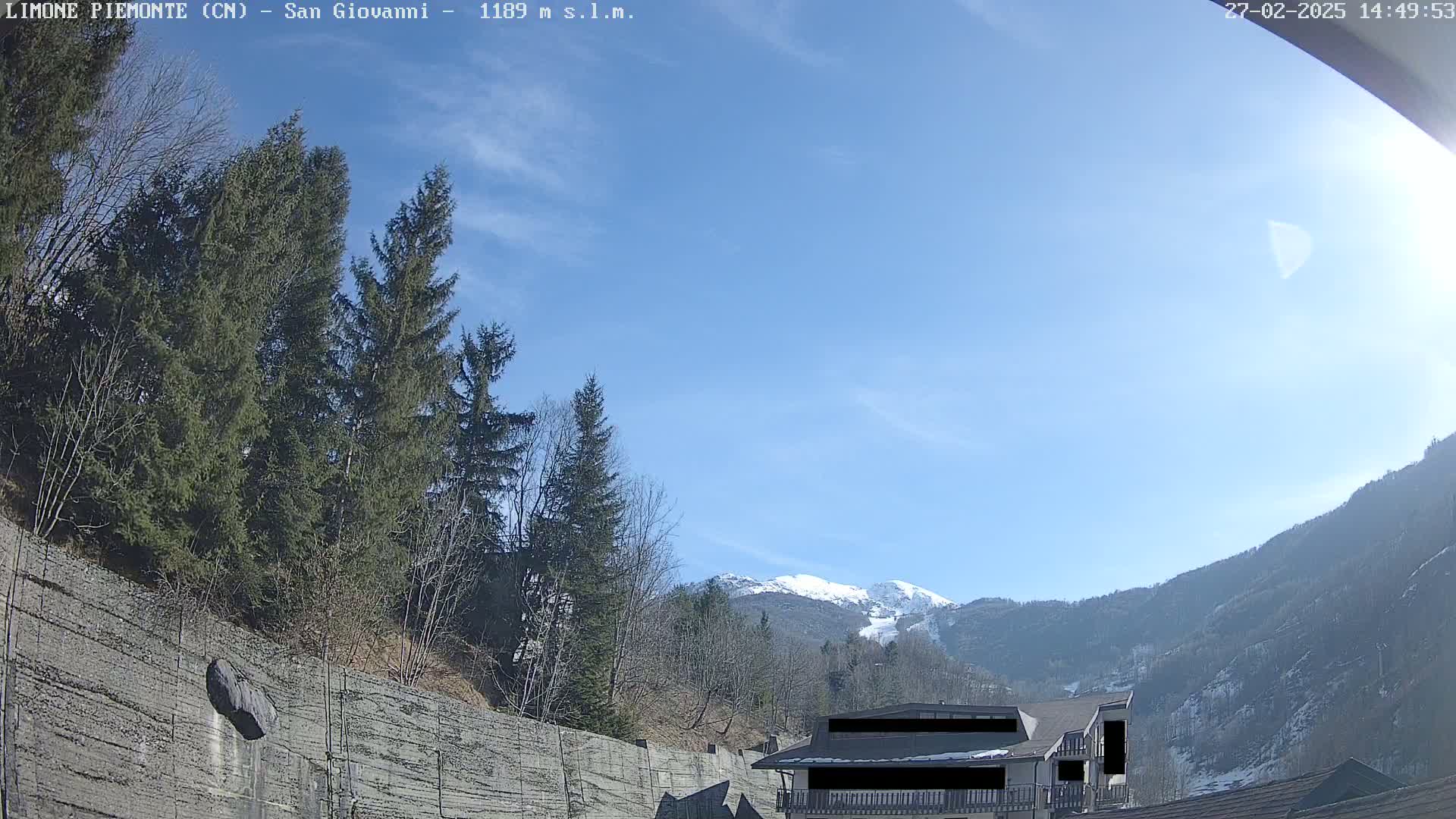 A sunny day in a mountainous region shows evergreen trees, a stone wall, and a building partially obscured, with snow-capped mountains in the background.