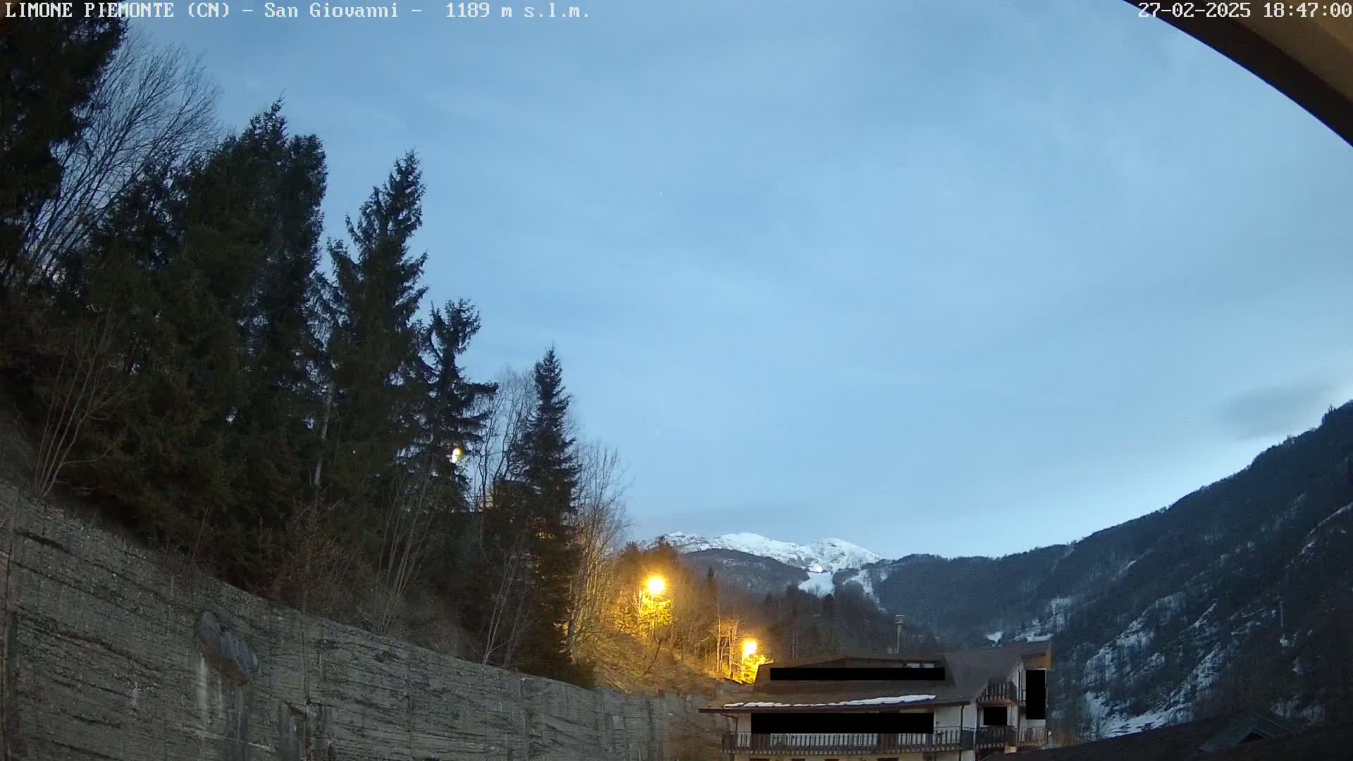 A twilight view shows evergreen trees and a rocky embankment next to a building, with snow-capped mountains in the distance under a clear, pale blue sky.