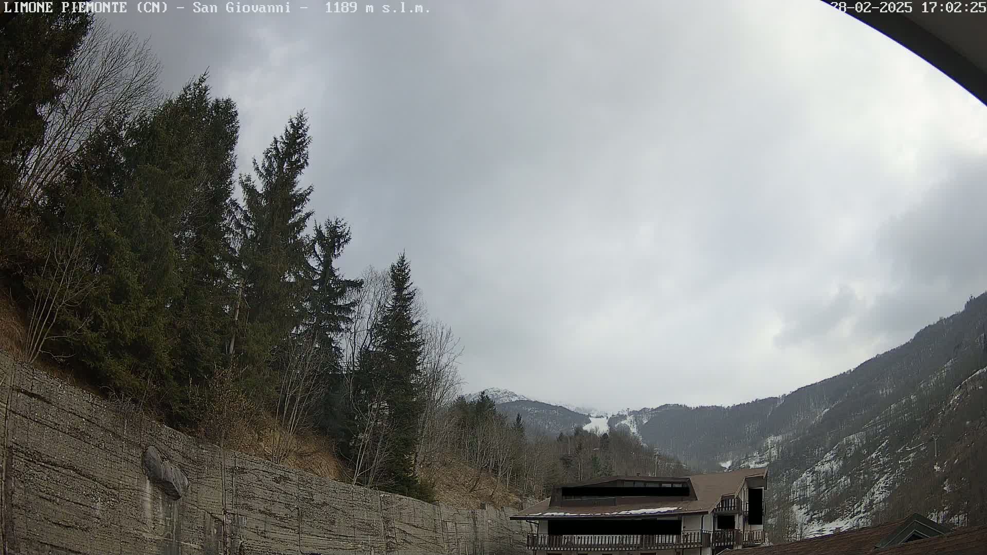 A cloudy day in a mountainous region shows evergreen trees and a building partially obscured by a retaining wall, with snow visible on distant hillsides.