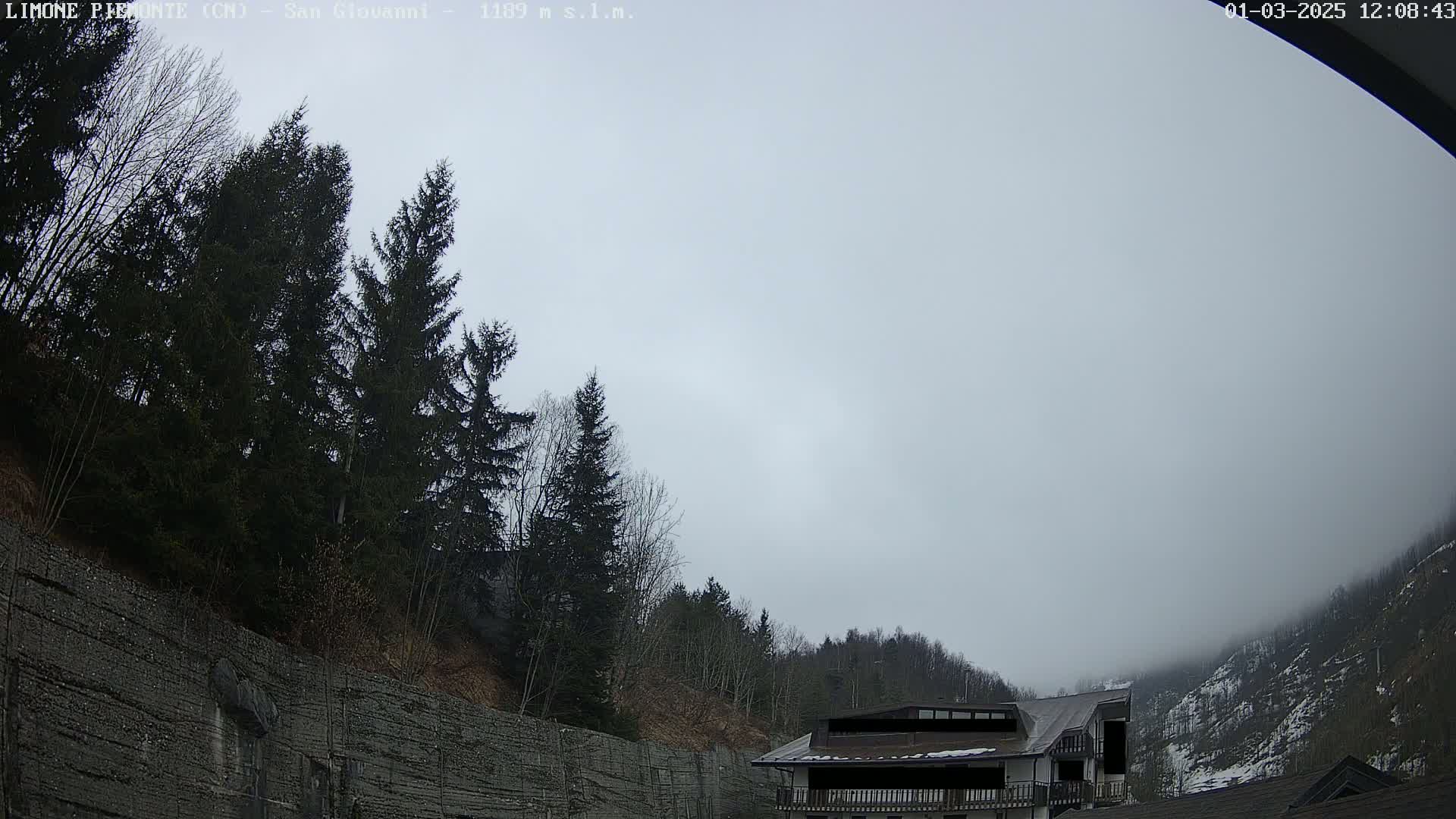 A mostly cloudy, overcast day reveals a mountainside with evergreen trees, a stone retaining wall, and a building partially obscured by fog.