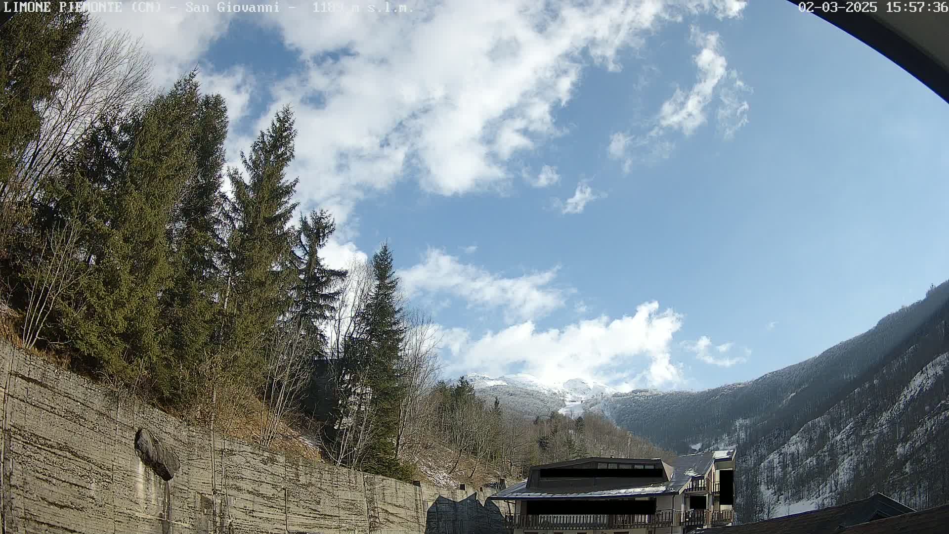 A partly cloudy sky overlooks a snow-dusted mountain range and evergreen trees behind a low stone wall.
