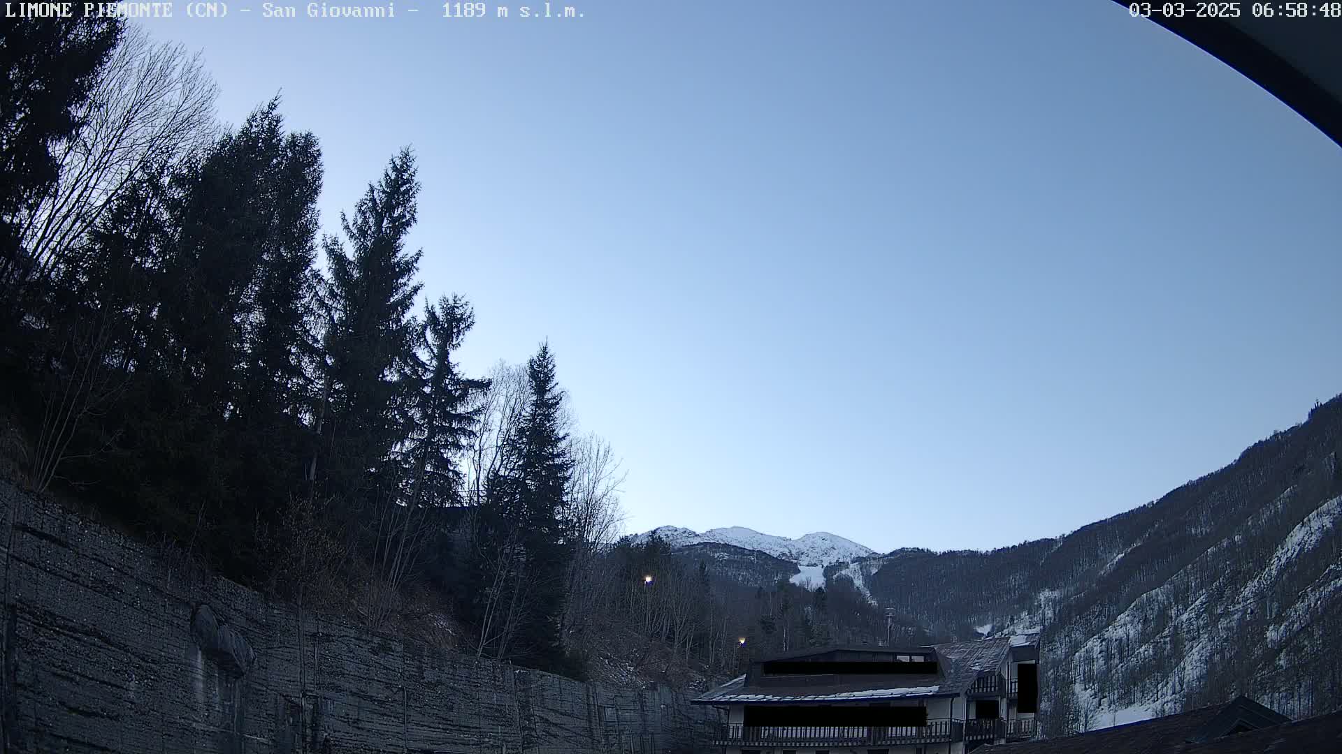 A clear, light-blue sky is visible above a mountainside with snow-dusted trees and a building partially obscured by the terrain.