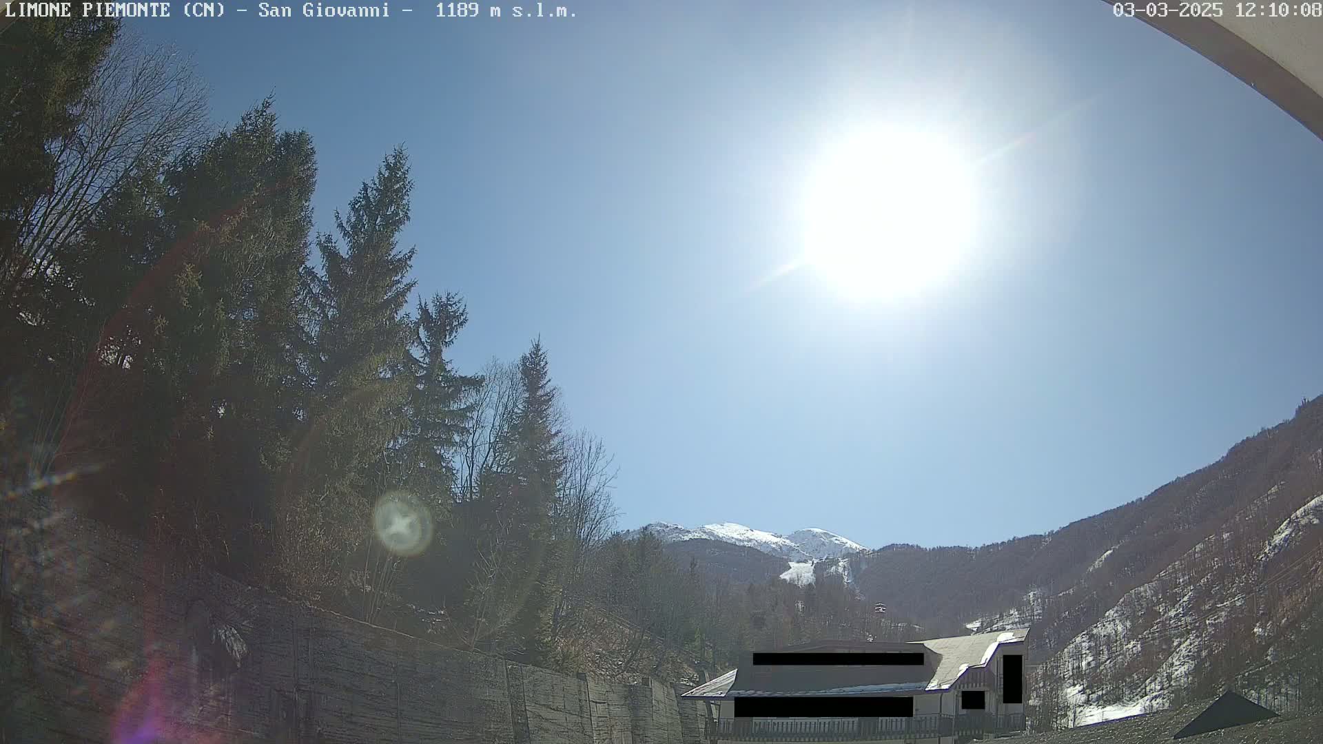 A sunny day in a mountainous area shows evergreen trees, a building partially obscured, and snow-capped mountains in the distance.
