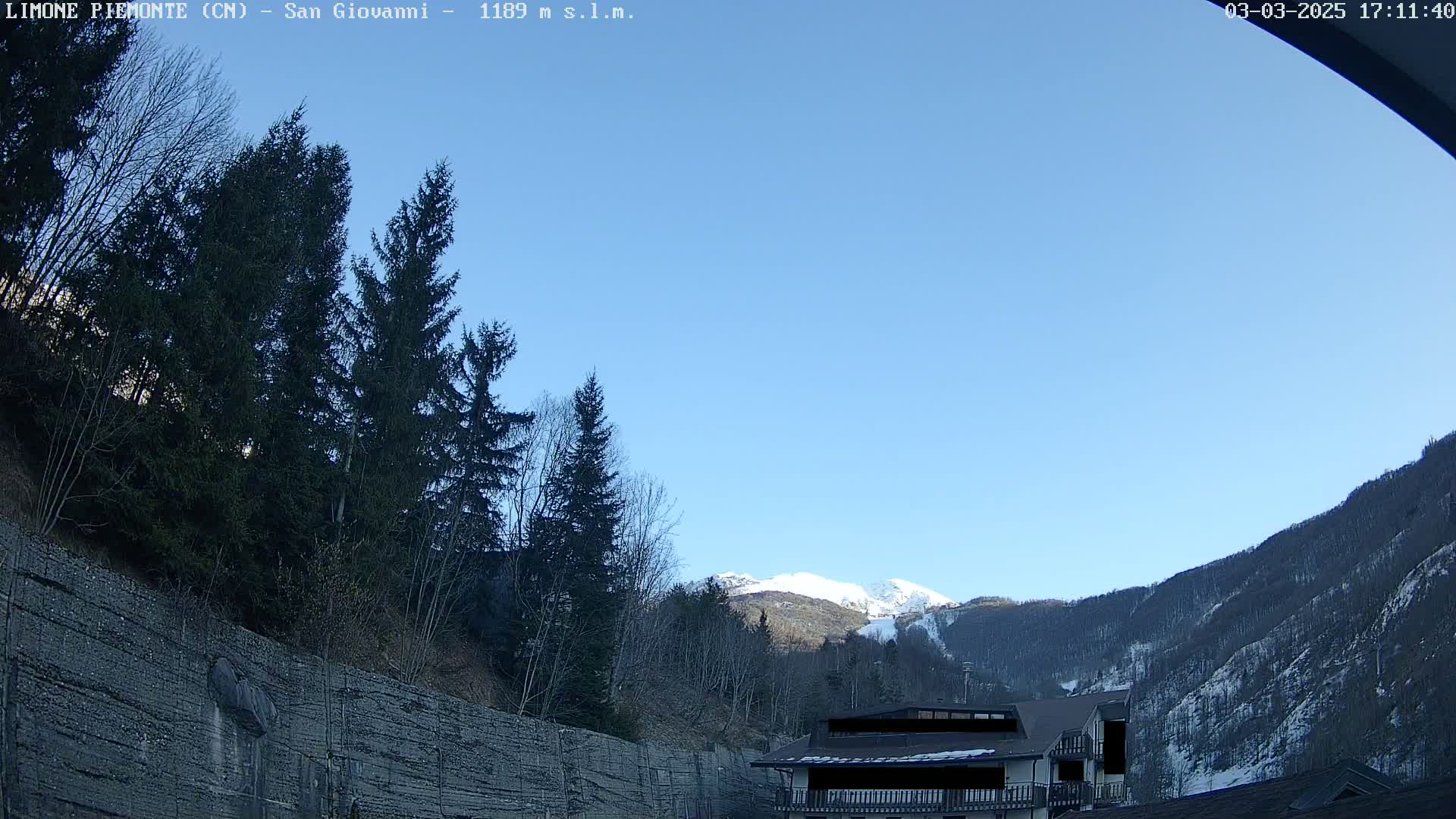 A clear blue sky is over snow-dusted mountains and evergreen trees, with a building partially visible in the foreground.