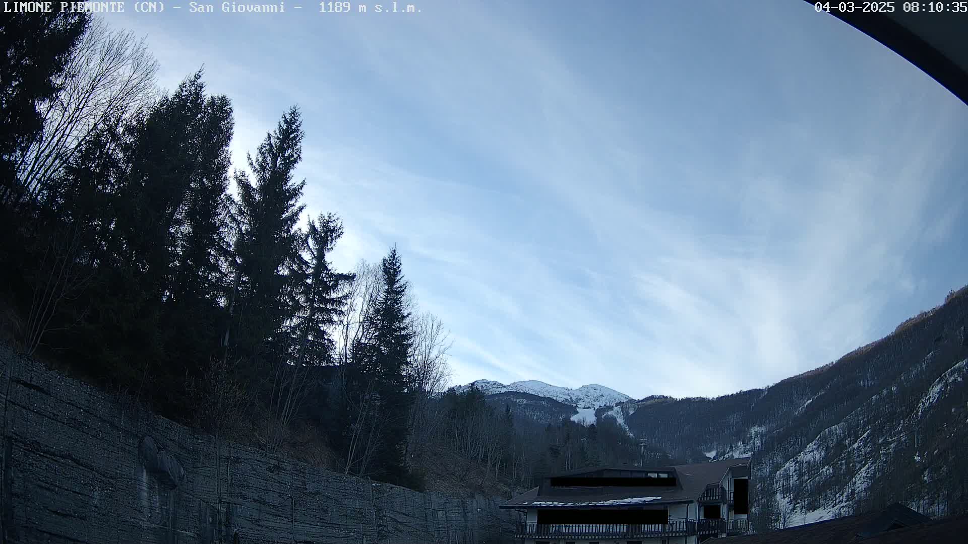 A mostly clear sky with wispy clouds overlooks a mountainside partially covered in snow, with dark evergreen trees in the foreground and a building partially visible near the bottom right.