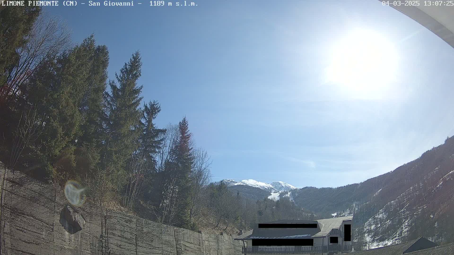 A sunny day in a mountainous region shows evergreens, a concrete wall, and a building partially obscured, with snow-capped mountains in the background.