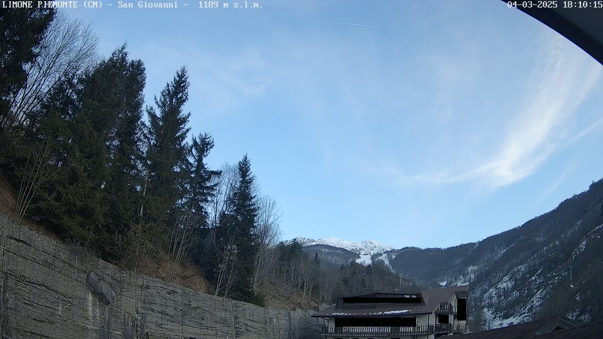 A mostly clear sky overlooks a snow-capped mountain behind a coniferous forest and a building, with a retaining wall in the foreground.