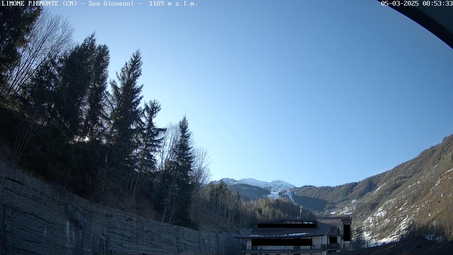 A clear, sunny day reveals a snow-capped mountain in the distance, partially obscured by trees and a building in the foreground.