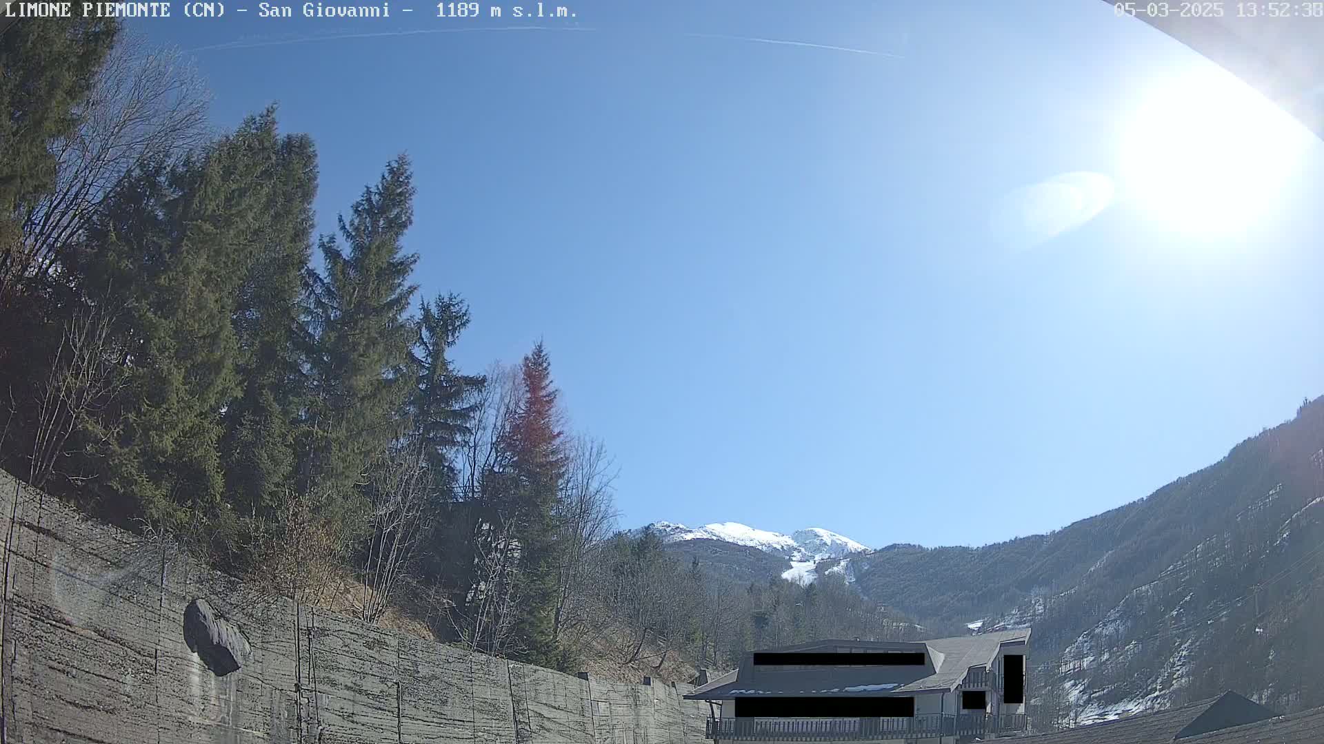 A sunny day reveals a snow-capped mountain in the distance, partially obscured by evergreens and a concrete retaining wall, with a building partially visible in the foreground.
