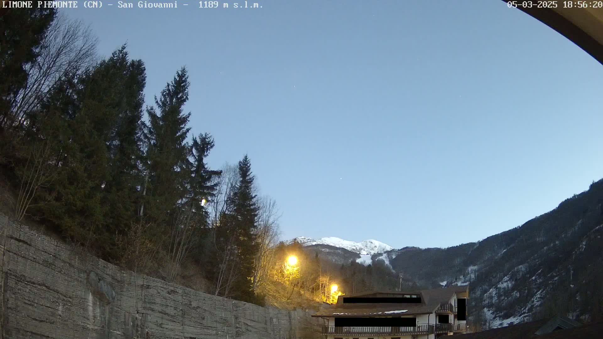 A twilight view shows a snow-capped mountain range behind a building nestled amongst dark evergreens on a hillside with a retaining wall in the foreground under a clear, pale blue sky.