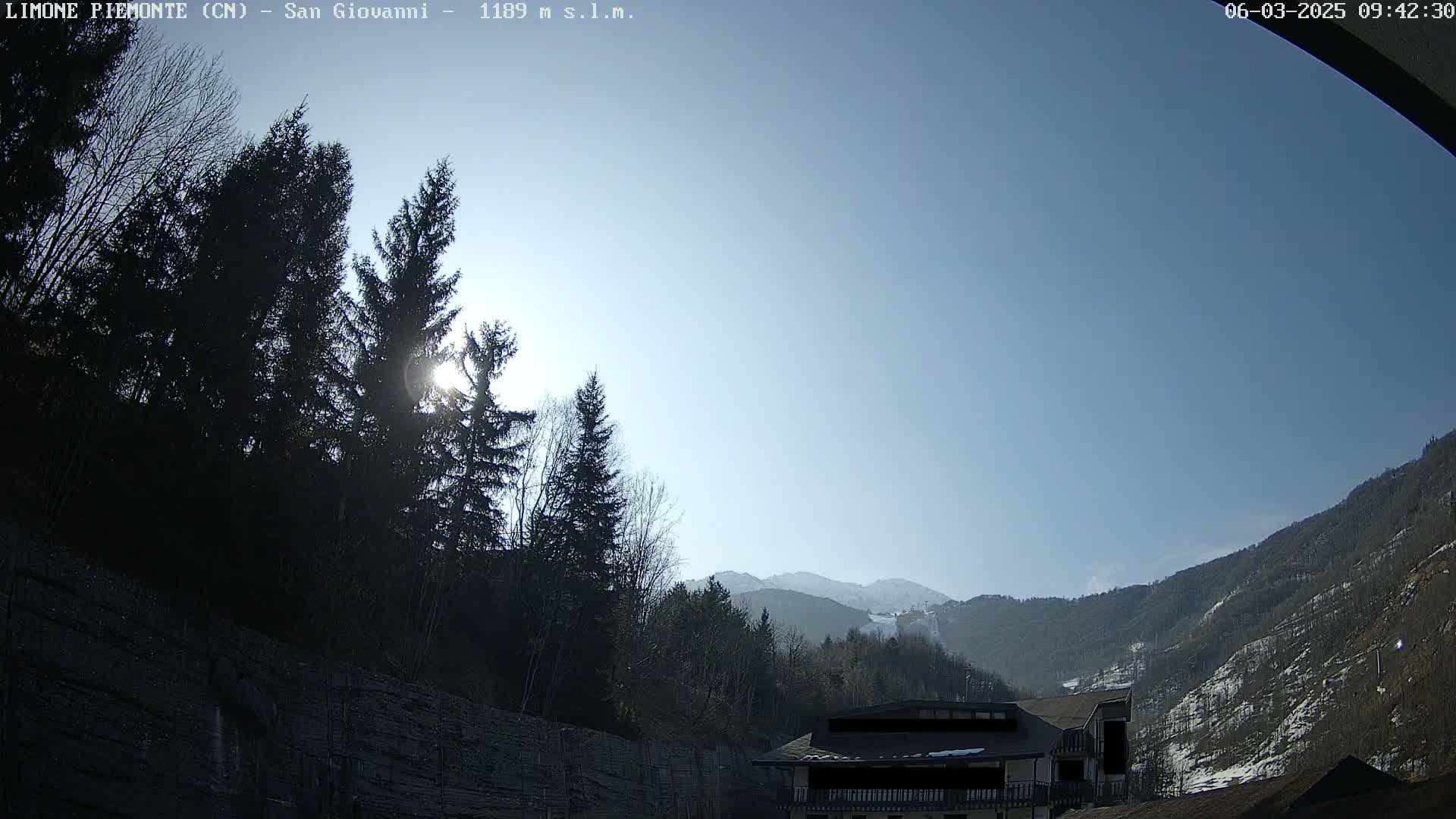 A sunny day reveals a mountain valley scene with evergreens in the foreground, a building partially visible, and snow-capped mountains in the distance.