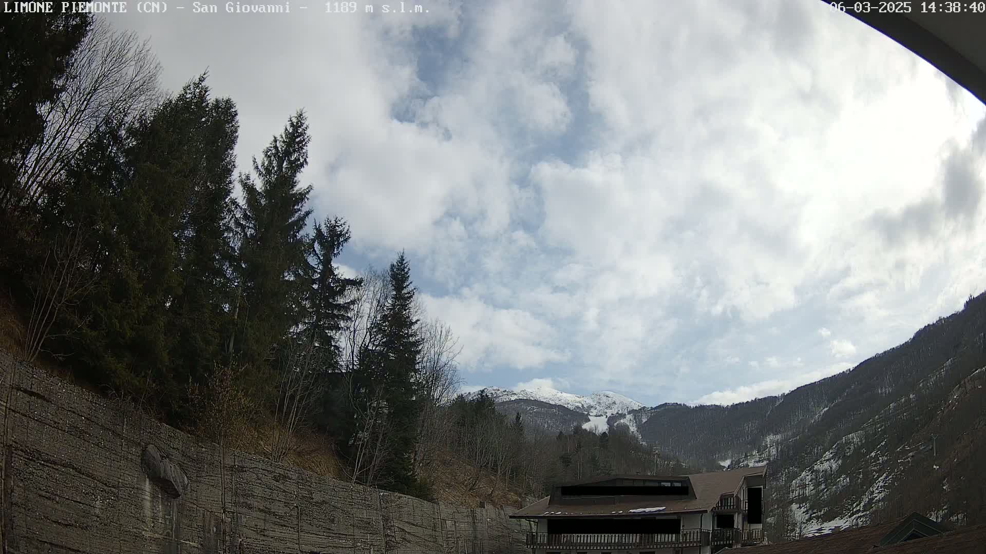 A partly cloudy day reveals snow-capped mountains in the distance, behind a line of evergreen trees and a building partially obscured by the camera angle.