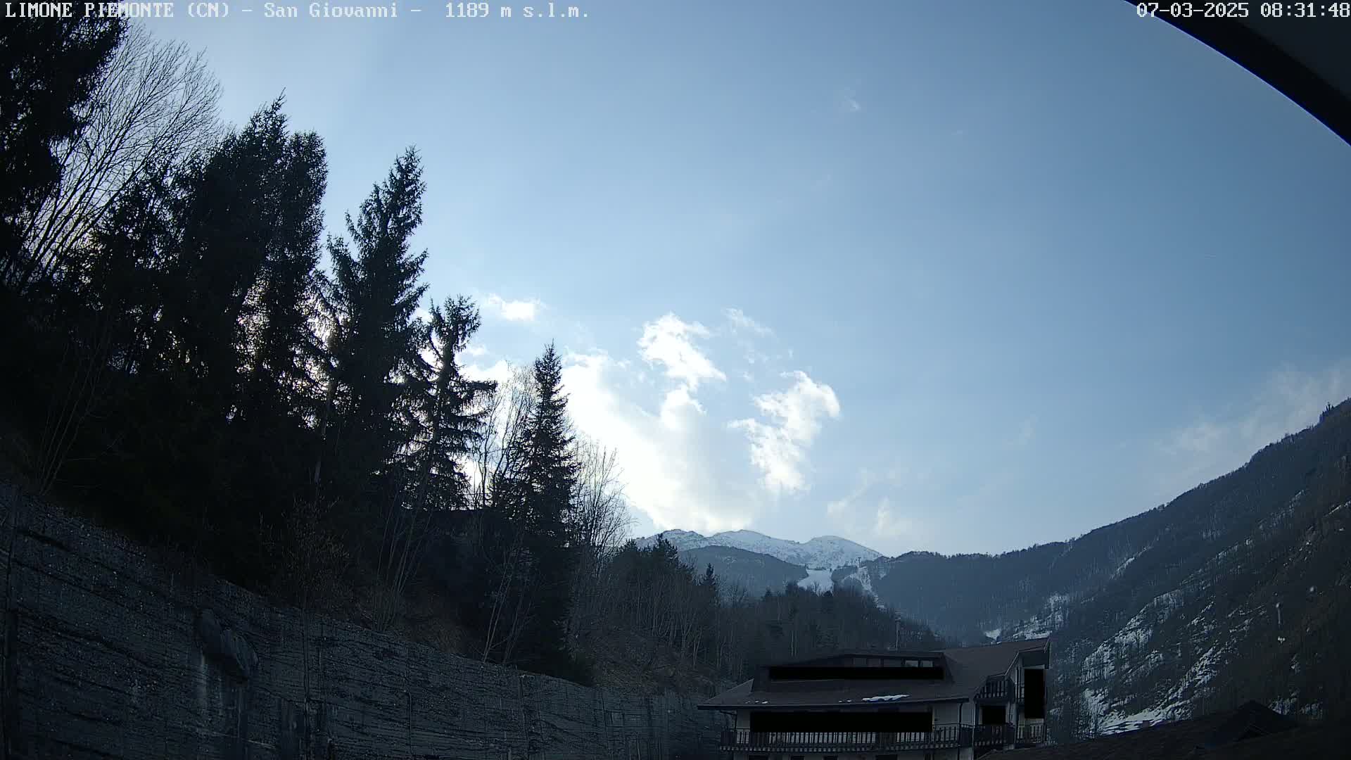 A partly cloudy sky overlooks a mountainside with dark evergreen trees, a building, and a snow-dusted mountain peak in the distance.