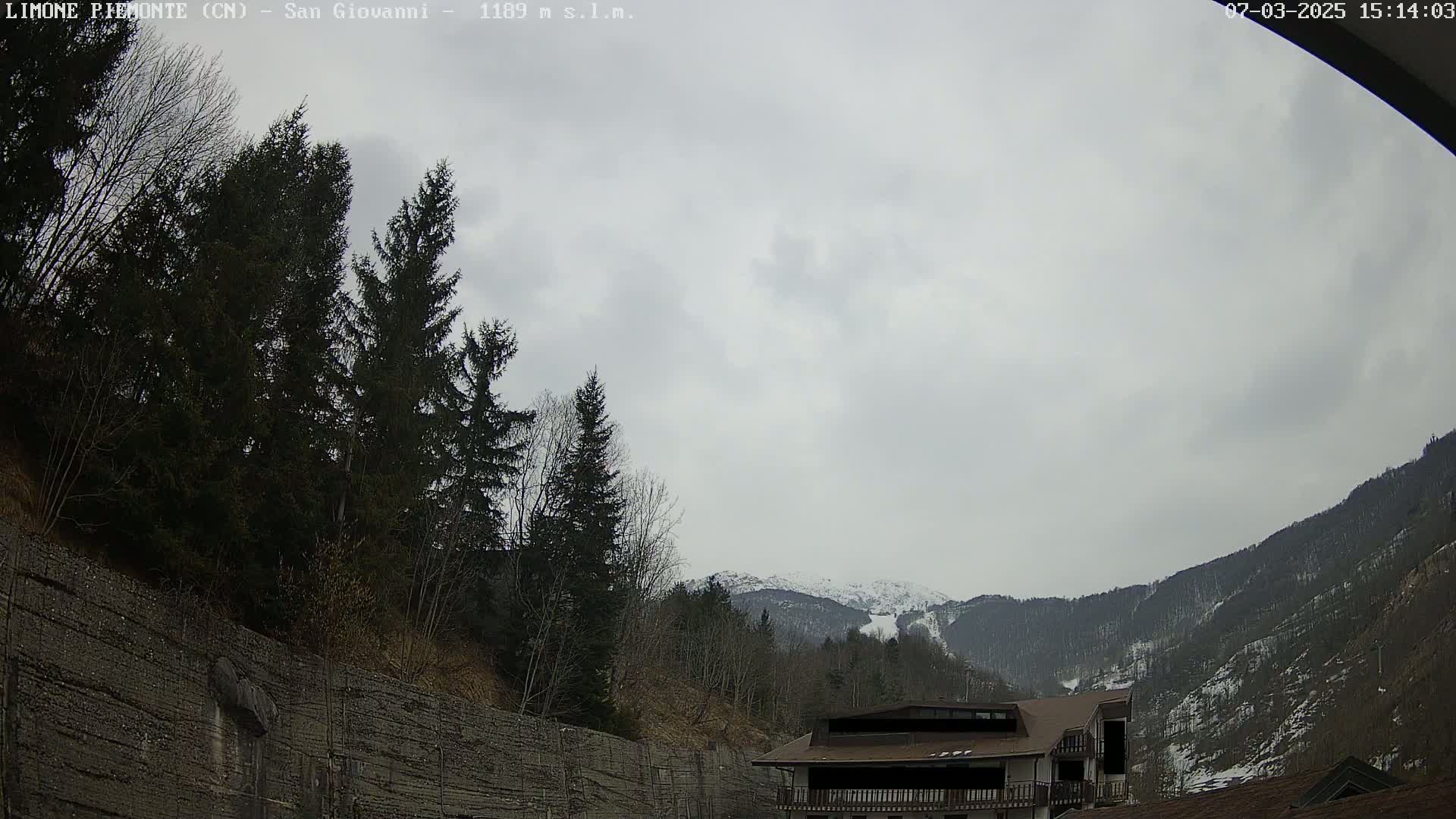 A cloudy day reveals a snow-dusted mountain range behind a building and a retaining wall partially covered in trees.