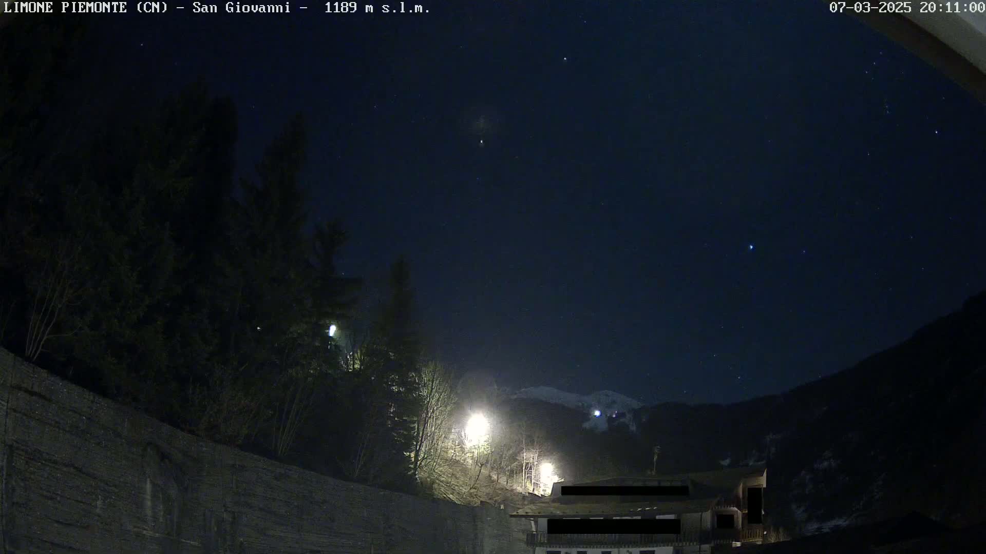A nighttime view of a dark mountain range under a clear, starry sky, illuminated by a few streetlights near a building partially obscured by shadows.