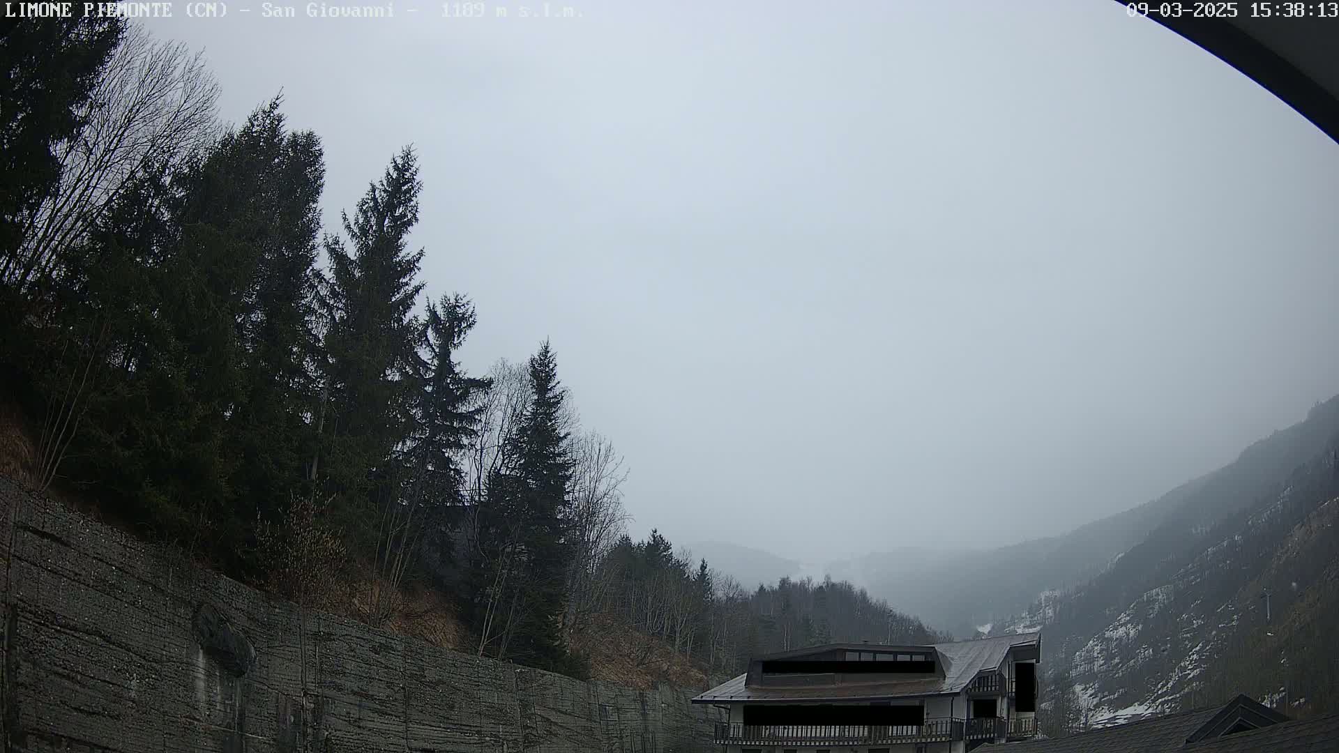 A foggy mountain valley is seen from a high vantage point, with evergreens and a building visible in the foreground.