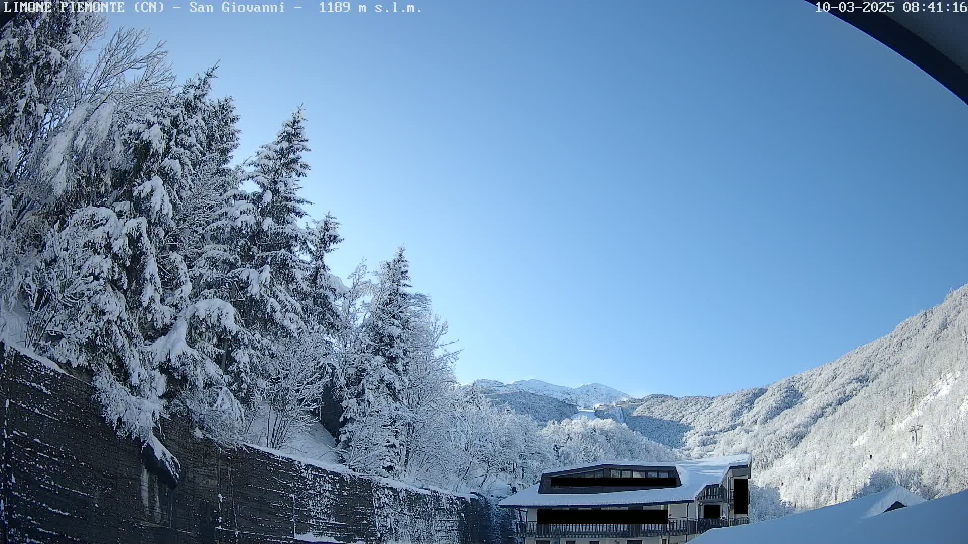 Snow-covered trees and a building sit on a mountainside under a clear blue sky.