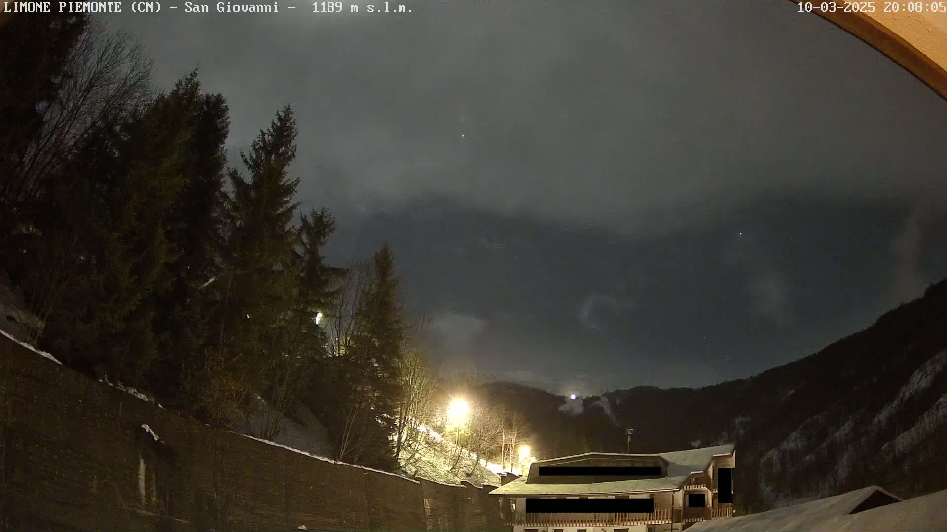 A nighttime view of a snow-covered mountainside with evergreen trees and a building, under a dark, cloudy sky.