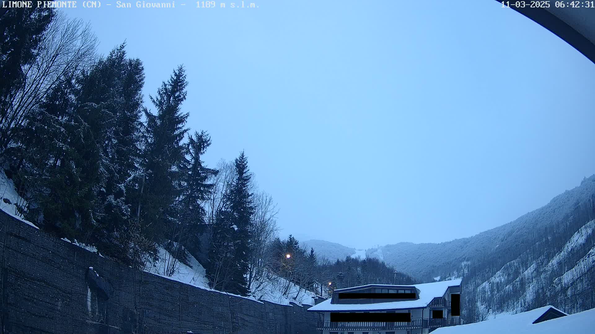 A snow-covered mountain landscape at dawn is seen, with evergreen trees in the foreground and a building nestled in the valley.