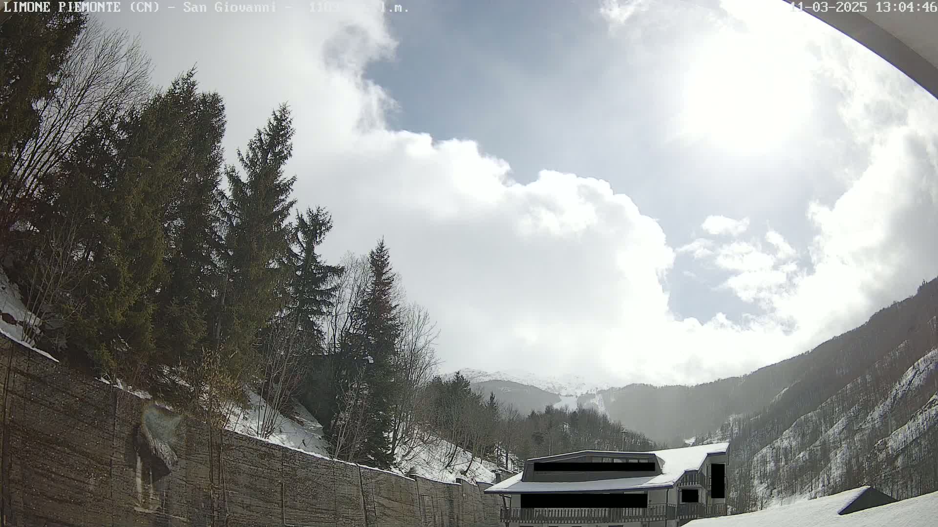 A partly sunny day with some clouds shows a snow-covered mountain landscape, featuring evergreen trees, a building, and a stone wall.