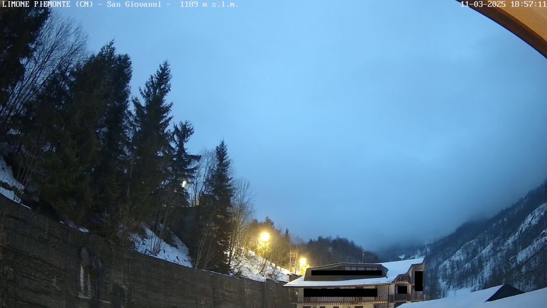 A twilight view of a snow-dusted mountainside with evergreens, a building, and a light fog or haze in the distance.
