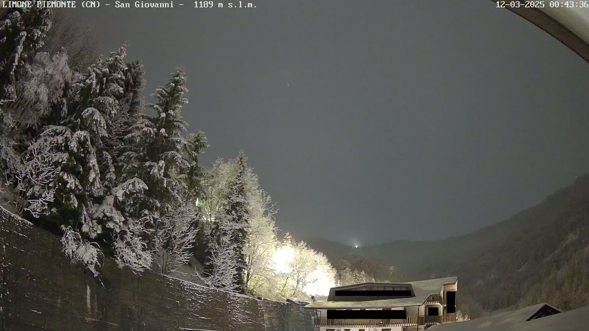 A snow-covered landscape at night shows a building nestled among snow-laden trees and mountains under a dark, cloudy sky.