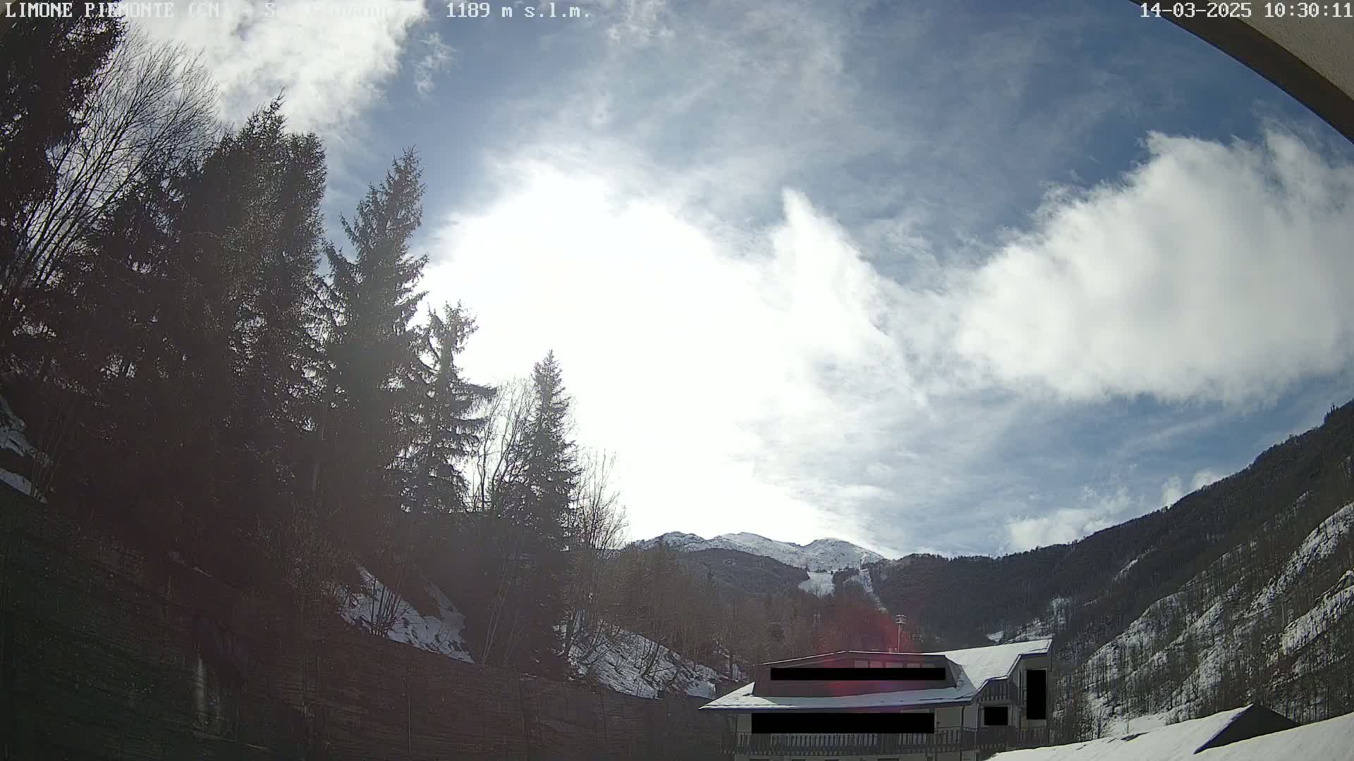 A partly cloudy sky brightens a snowy mountainside scene, where evergreens stand beside a house partially obscured by snow.