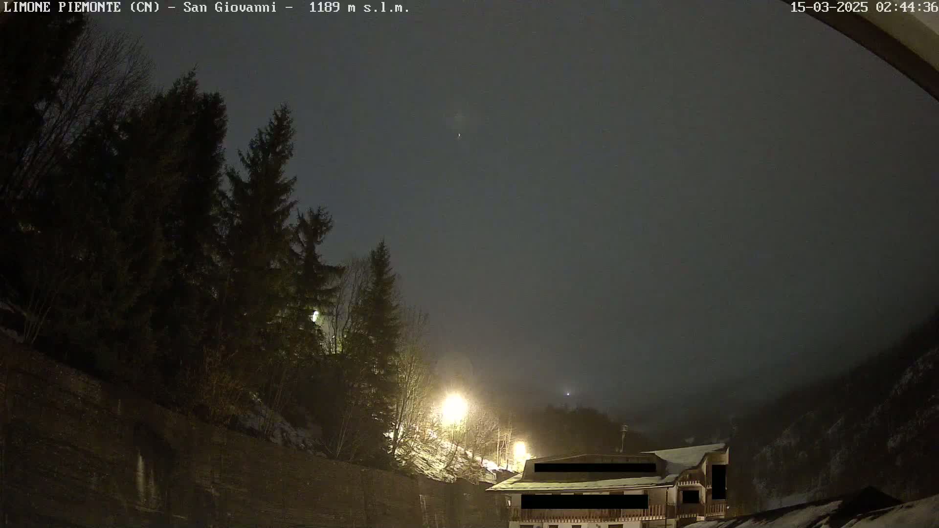 A nighttime scene shows a building partially obscured by fog and snow, nestled among dark evergreens under a dark, overcast sky.