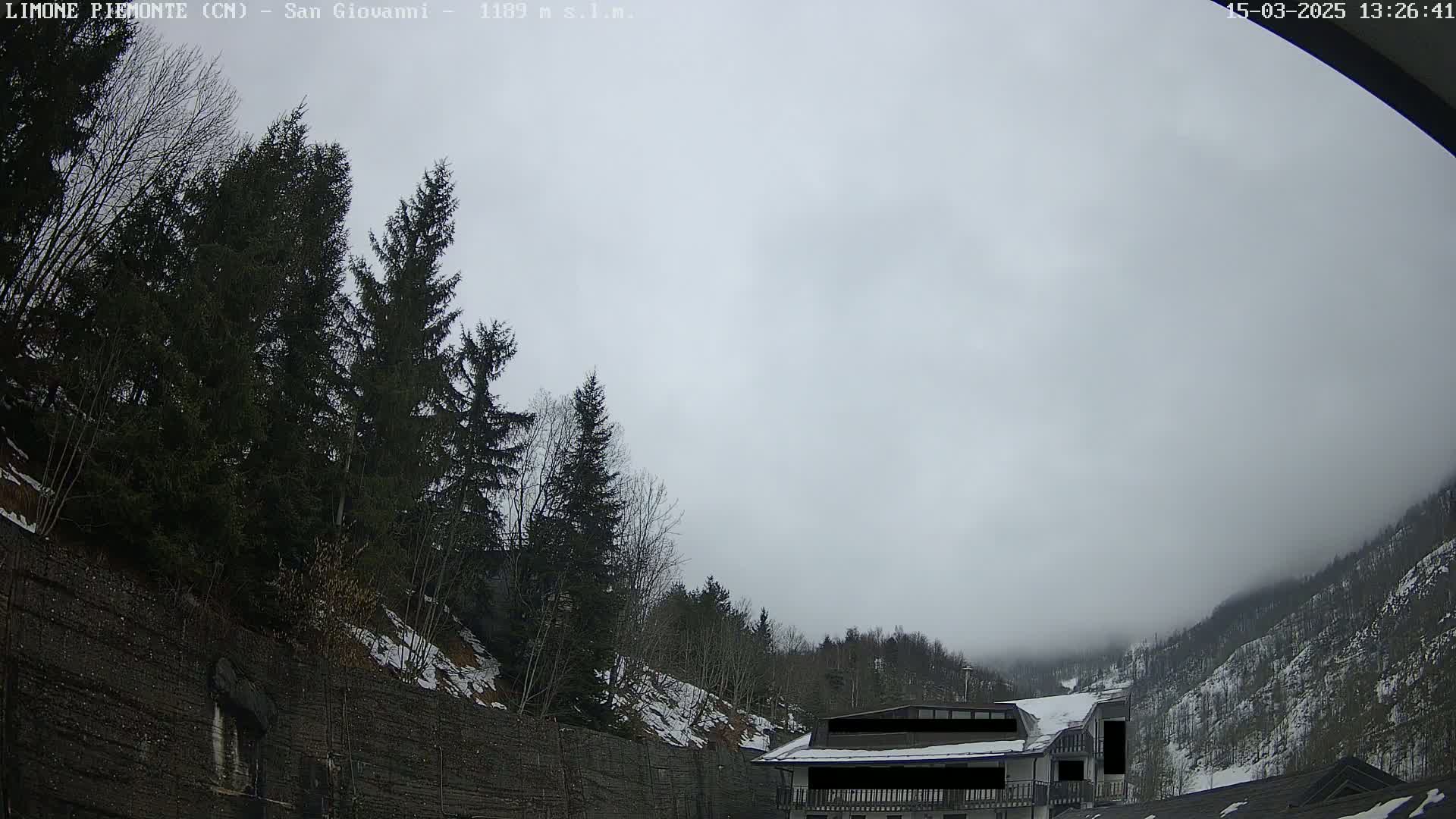A mostly cloudy, possibly foggy day reveals a mountainside with evergreen trees, a building partially obscured by fog, and a retaining wall with patches of snow.