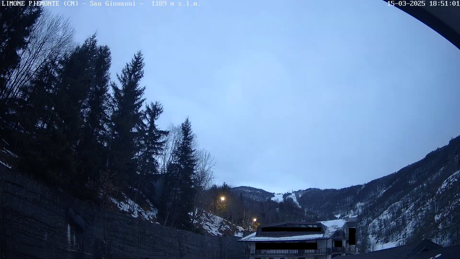 A clear twilight sky is visible above a snow-dusted mountainside with dark evergreen trees and a building partially obscured.