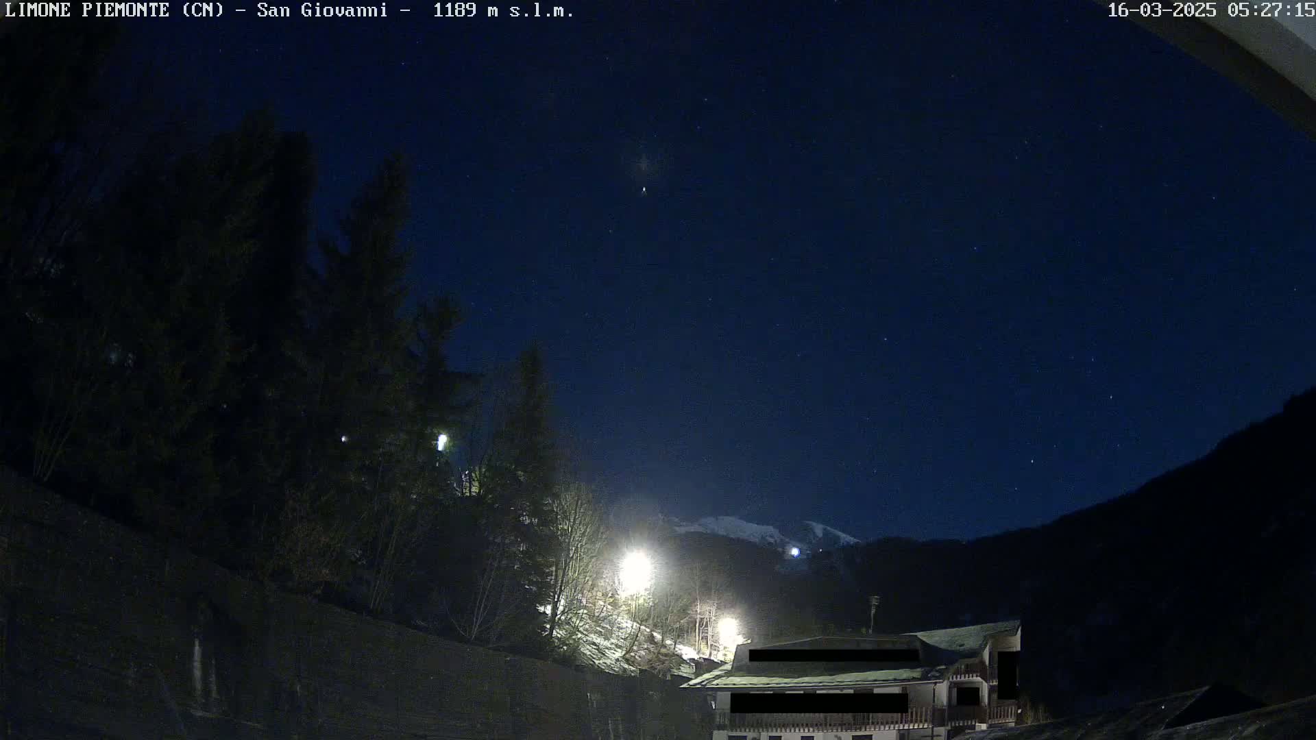 A nighttime view of a snow-covered mountain behind dark trees and a building, under a clear, starry sky.