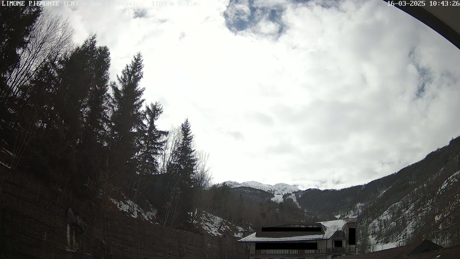 A cloudy sky hangs over a mountainous landscape with dark evergreen trees, a building partially obscured, and patches of snow visible on the mountainside and ground.