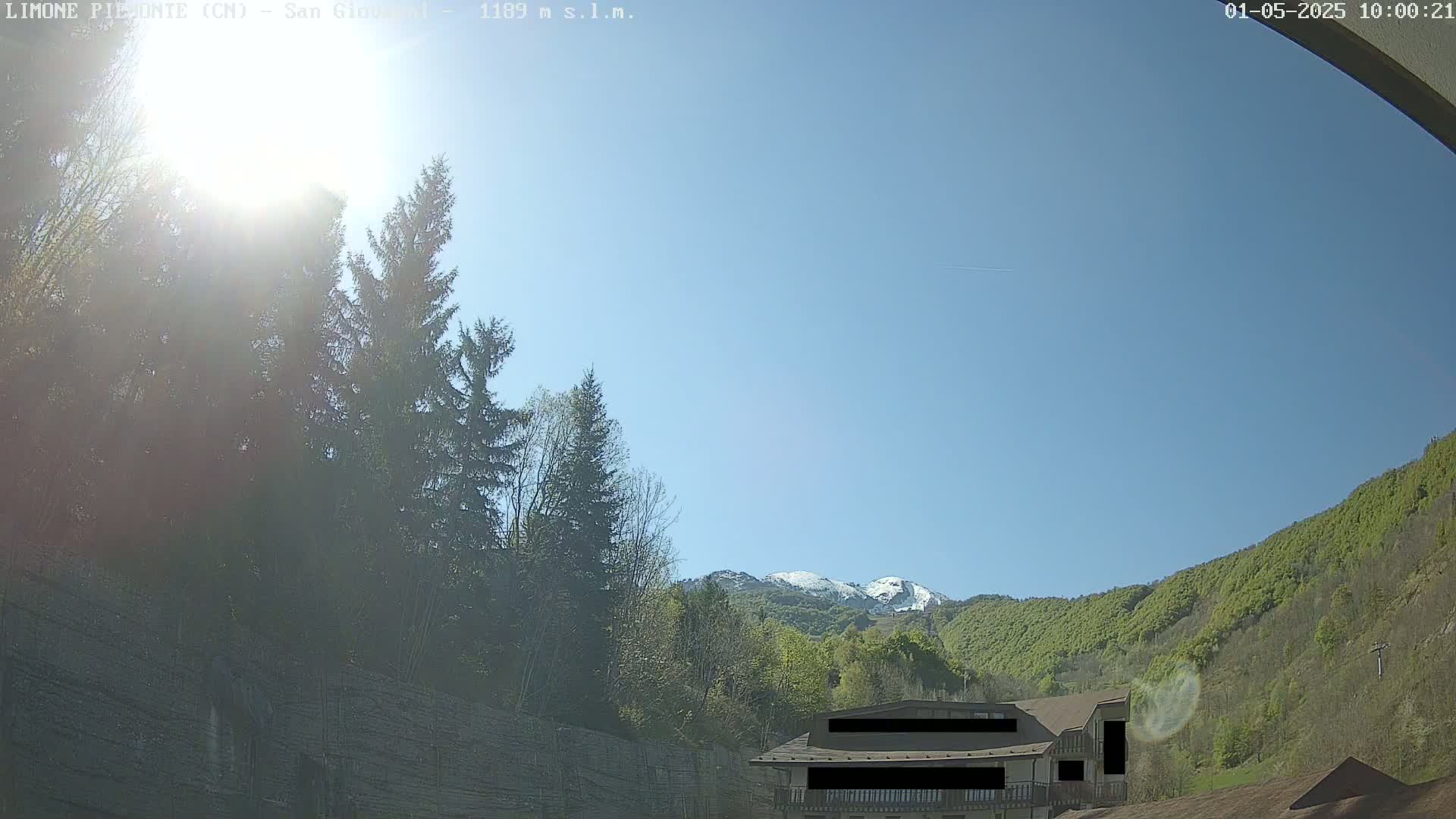 A sunny day reveals a snow-capped mountain in the distance, partially obscured by a lush green hillside and a building partially visible amidst evergreen trees.