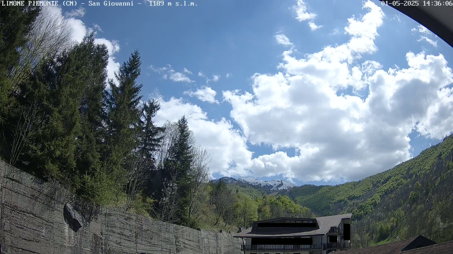 A partly cloudy sky overlooks a mountainside with lush green trees and a building partially obscured by a concrete retaining wall.