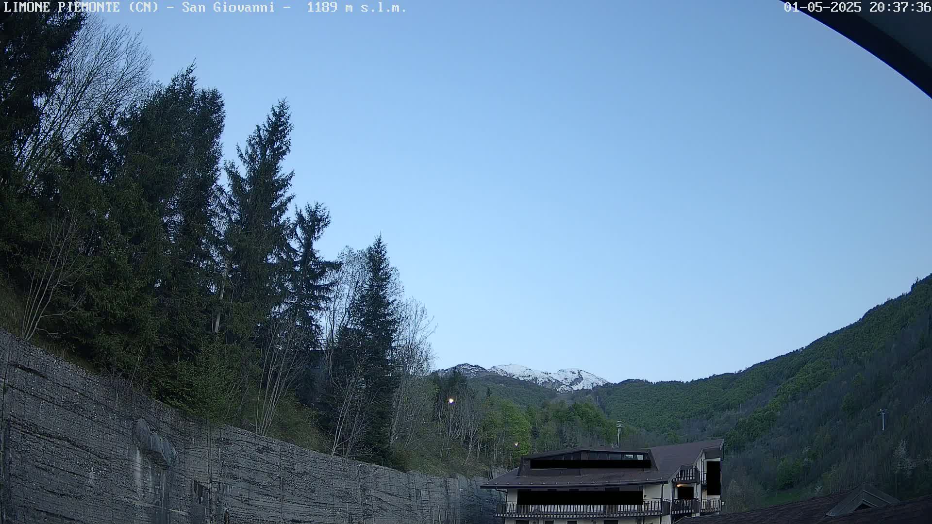 A clear, twilight sky overlooks a mountain range partially covered in snow, with evergreen trees and a building nestled amongst the foothills.