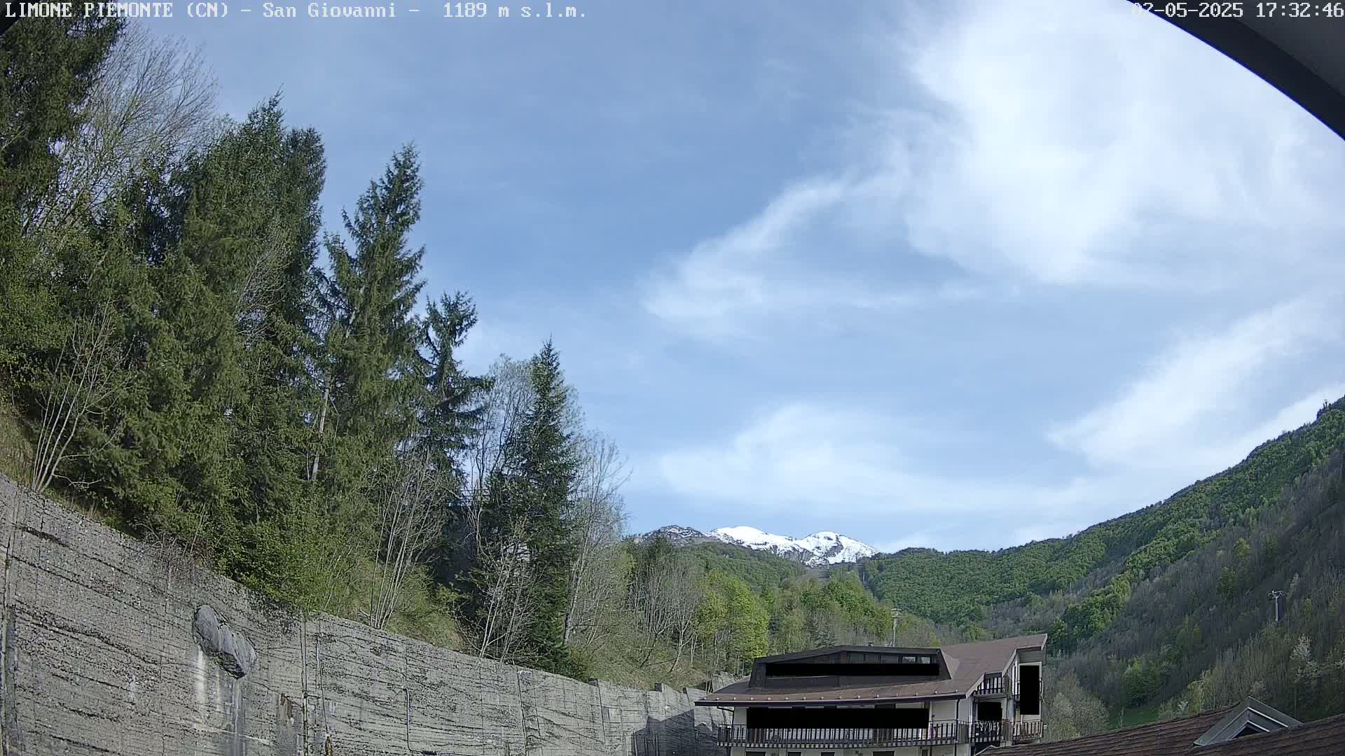 A partly cloudy sky overlooks a mountainside with lush green trees, a building, and a retaining wall.