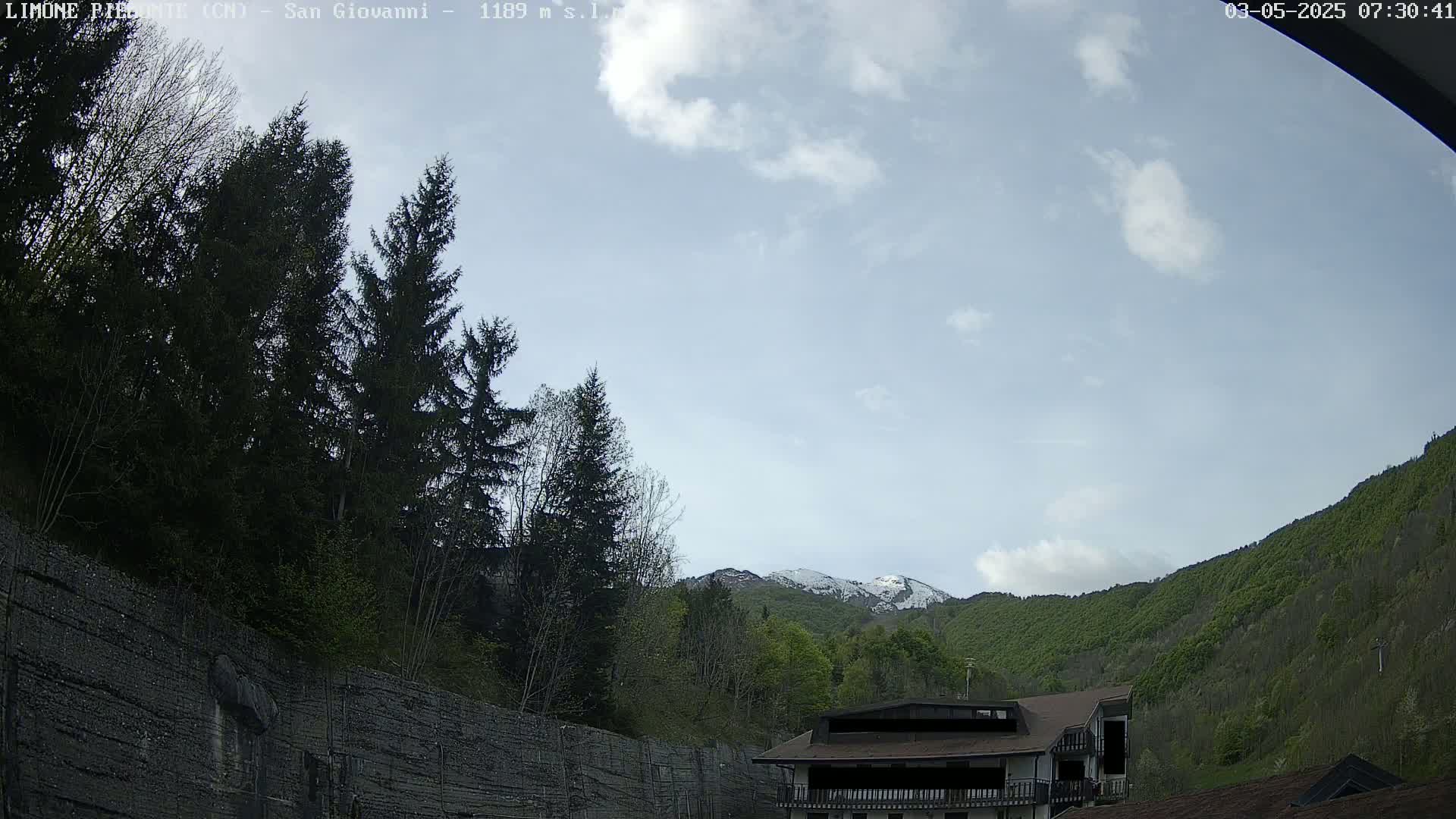 A partly cloudy sky overlooks a mountain range with patches of snow, a building nestled amongst green trees and a tall, dark retaining wall.