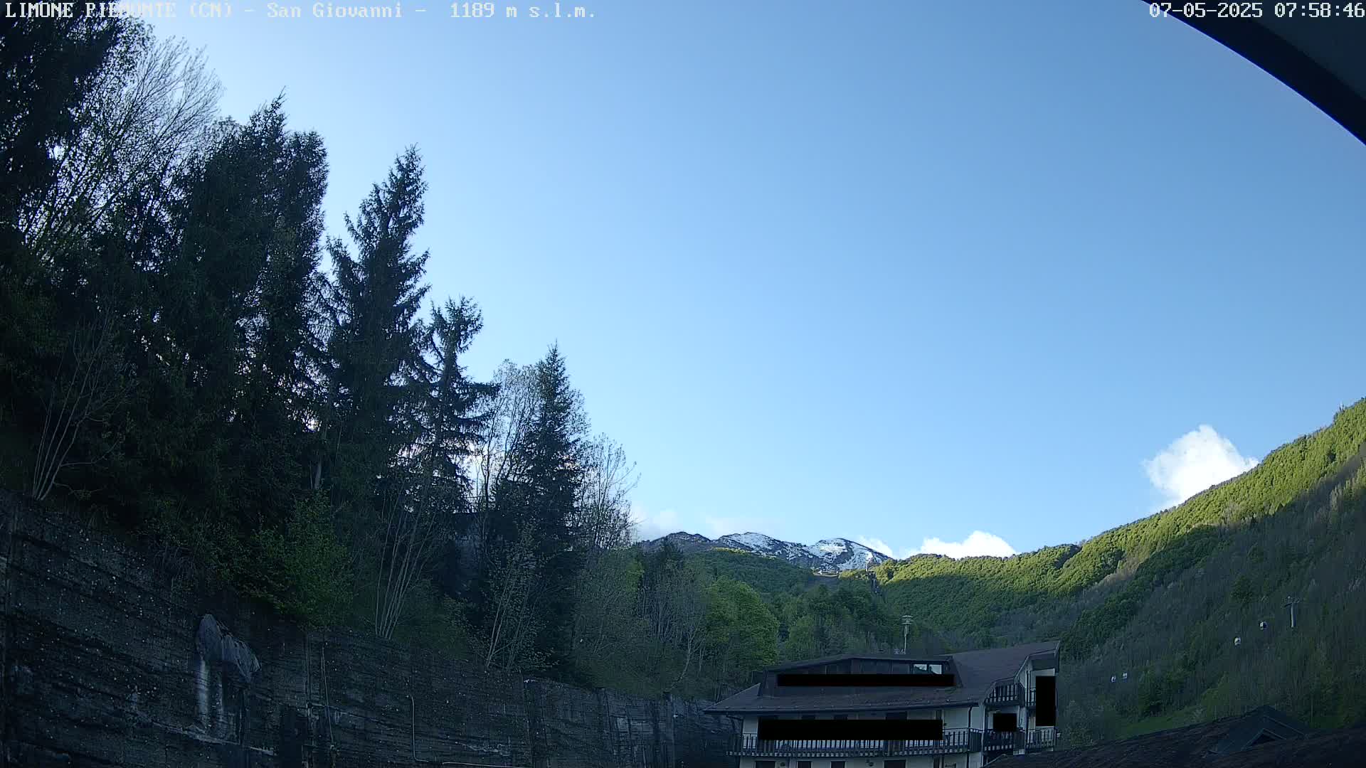 A mostly clear blue sky overlooks a mountainside with snow-capped peaks, lush green vegetation, and a building nestled amongst the trees.