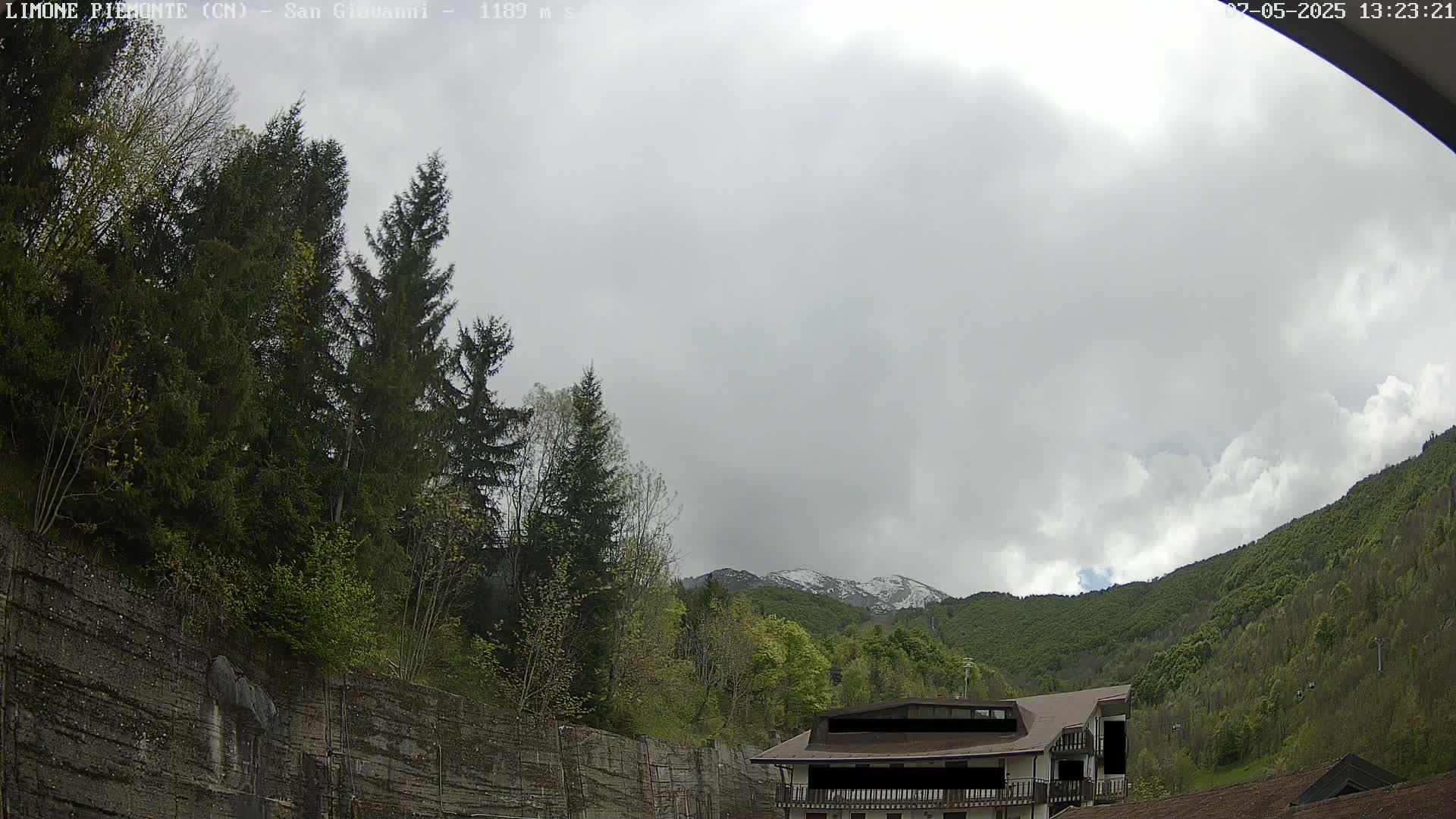 A cloudy day reveals a snow-dusted mountain behind a building nestled in a forest of evergreens and deciduous trees, partially obscured by a retaining wall.