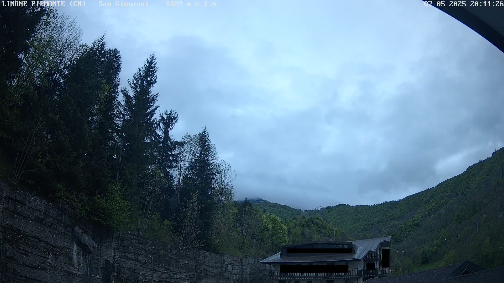 A cloudy sky hangs over a mountainside, where evergreens and deciduous trees grow alongside a stone wall and a building.