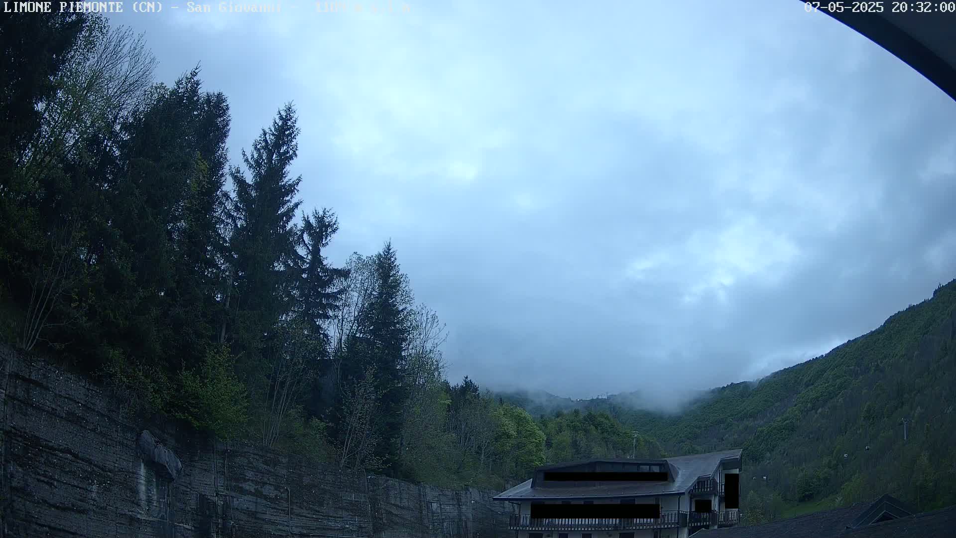 A cloudy evening scene shows a building nestled among evergreens on a hillside, with fog rolling in over the mountains in the distance.