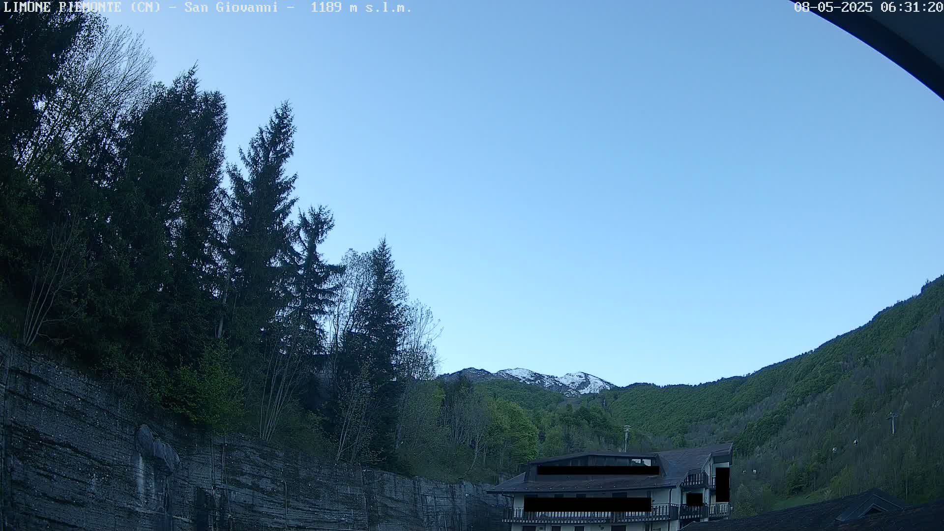 A clear blue sky overlooks a mountainside covered in green trees, with a partially snow-capped mountain peak in the distance and a building nestled amongst the trees.