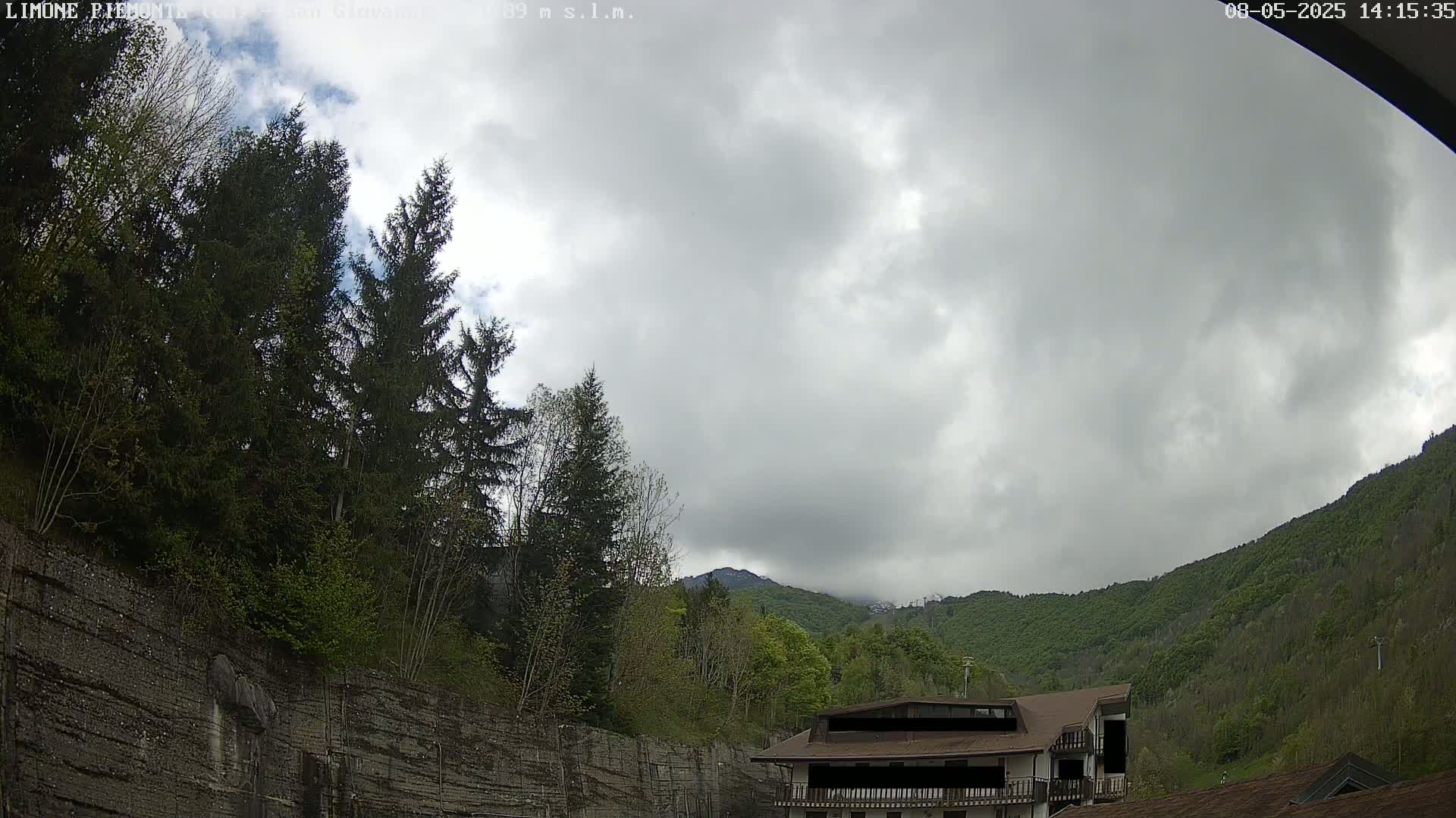 A cloudy sky hangs over a mountainside covered in lush green trees and a building nestled amongst the foliage.