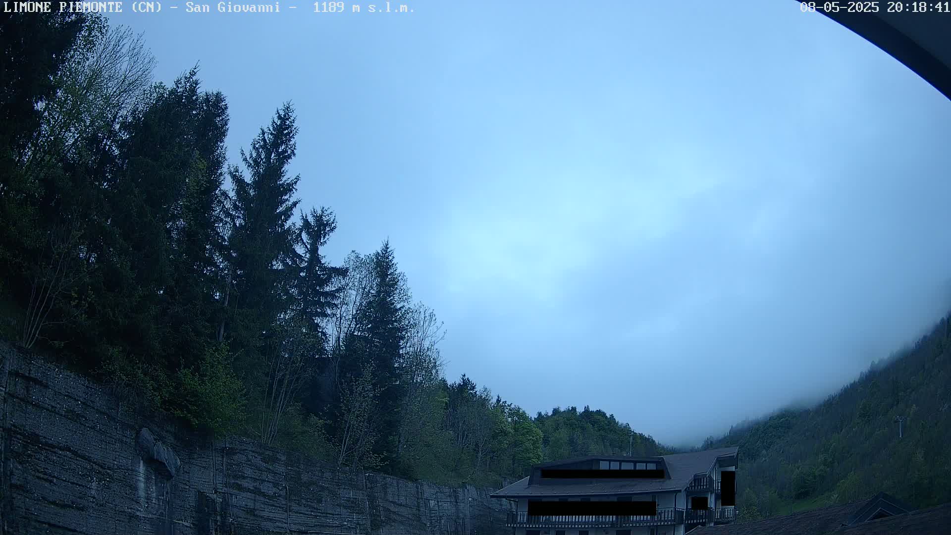 A hazy, light blue sky hangs over a mountainside where evergreen trees and a building are partially obscured by fog.