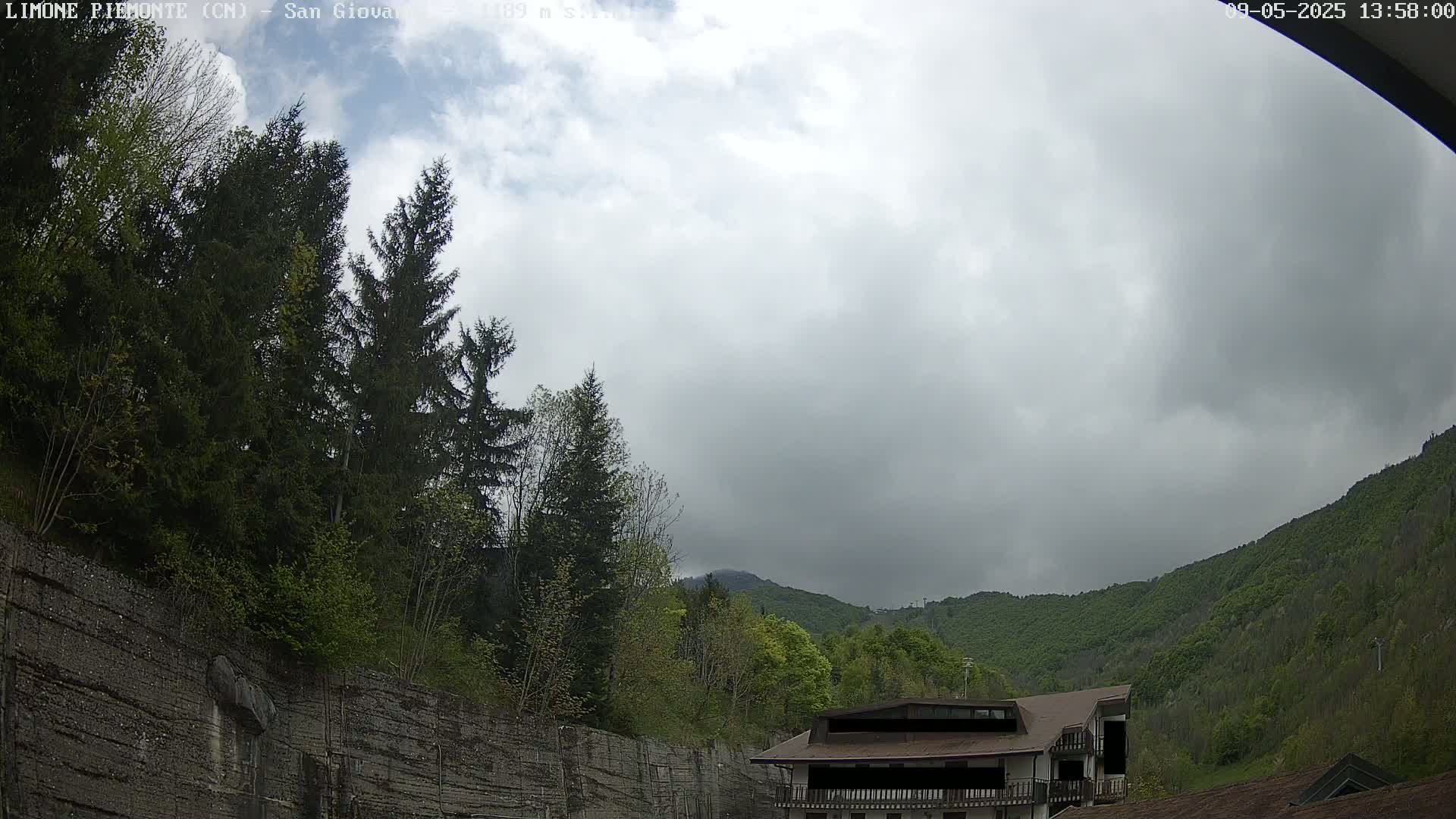 A cloudy day reveals a mountainside covered in lush green trees, partially obscured by a stone wall, with a building nestled among the foliage.