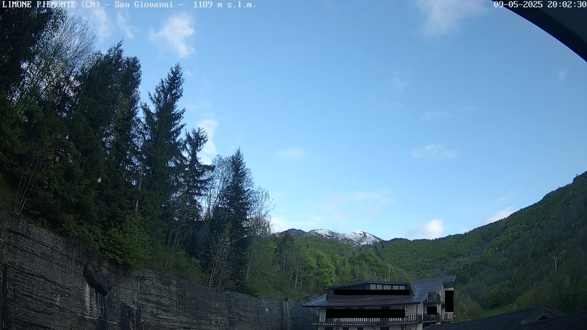 A mostly clear blue sky overlooks a mountainside with green trees, a snow-capped peak, and a building partially visible behind a stone wall.