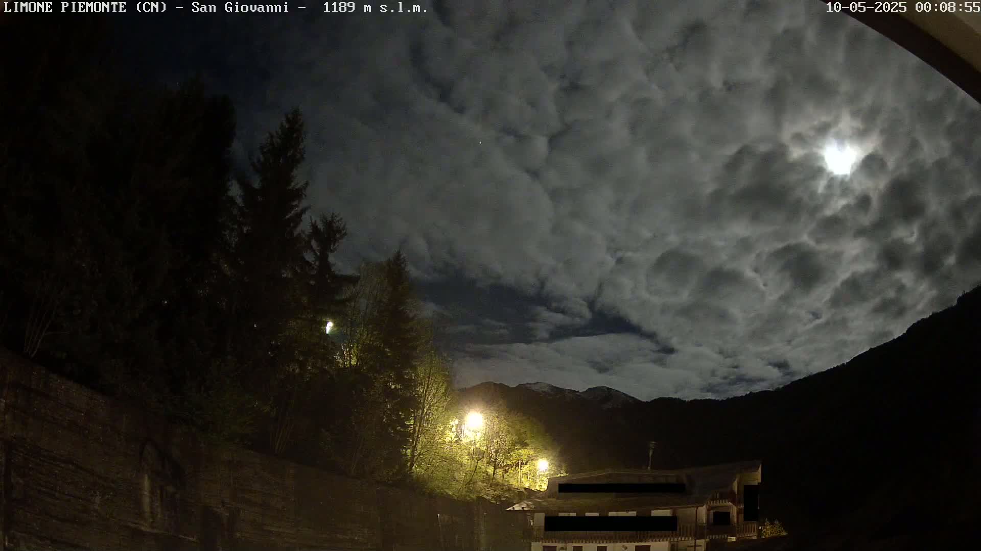A nighttime view of a mountainous area under a heavily clouded sky with a partially visible moon, illuminated by streetlights near dark evergreen trees and buildings.
